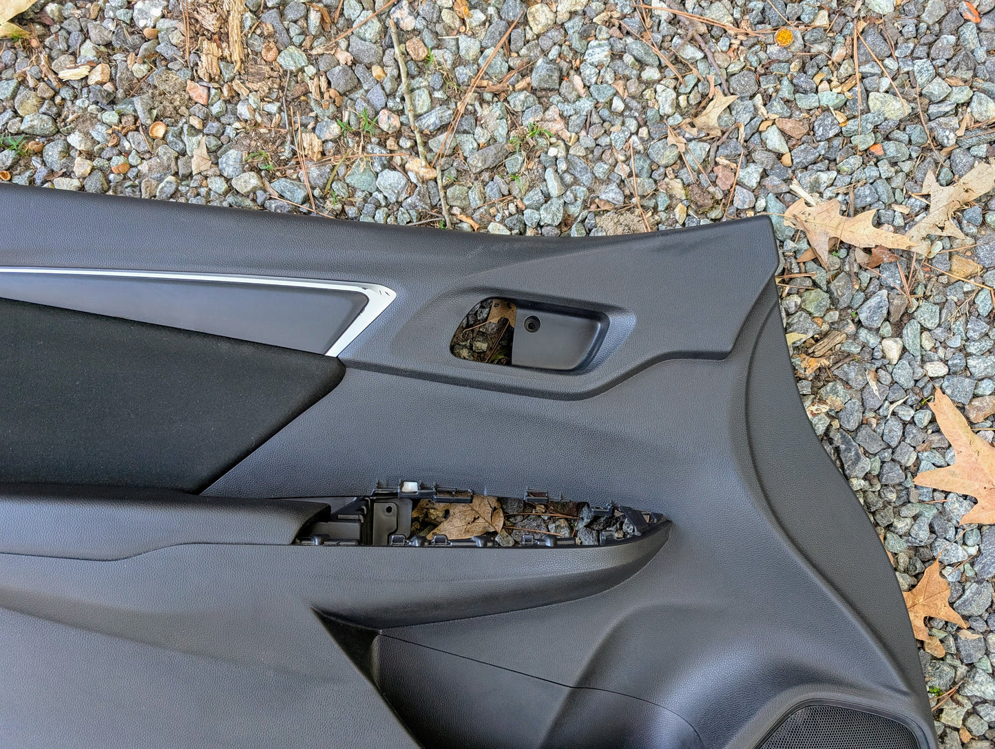 An OEM Honda Front Driver Power Door Panel for 2015-2020 Fit LX Trim is shown detached on gravel with brown leaves, missing the door handle and window switch.