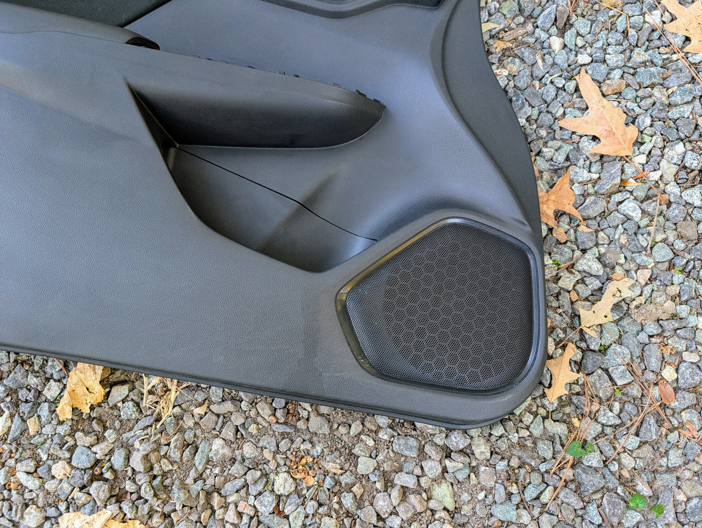A close-up of a Honda OEM Front Driver Power Door Panel for 2015-2020 Fit LX Trim, showing a hexagonal speaker grille, seen lying on gravel with scattered brown leaves.