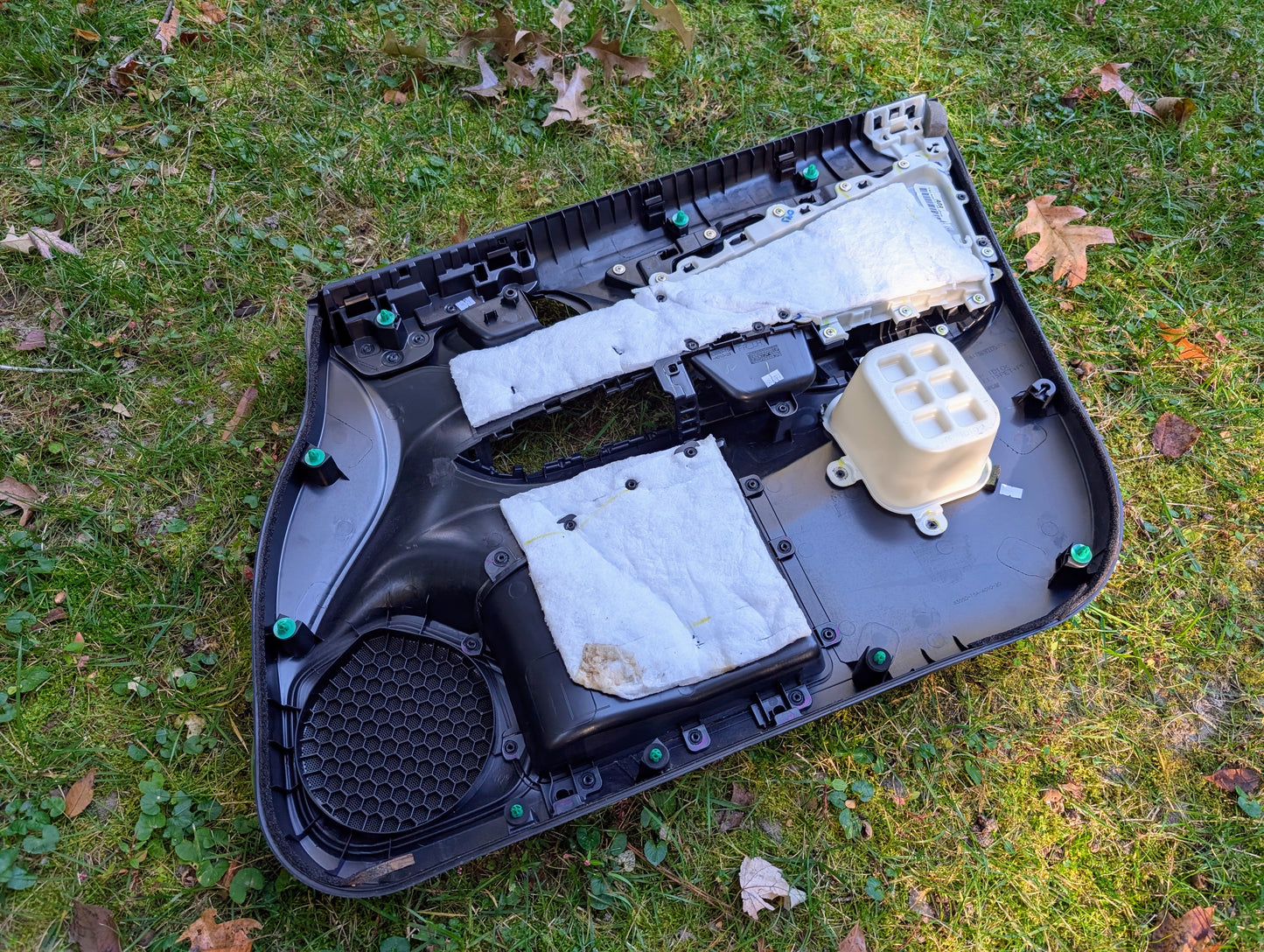 A black OEM Front Driver Power Door Panel for 2015-2020 Honda Fit LX by Honda, featuring white insulation and fasteners, rests on grass with scattered brown leaves.