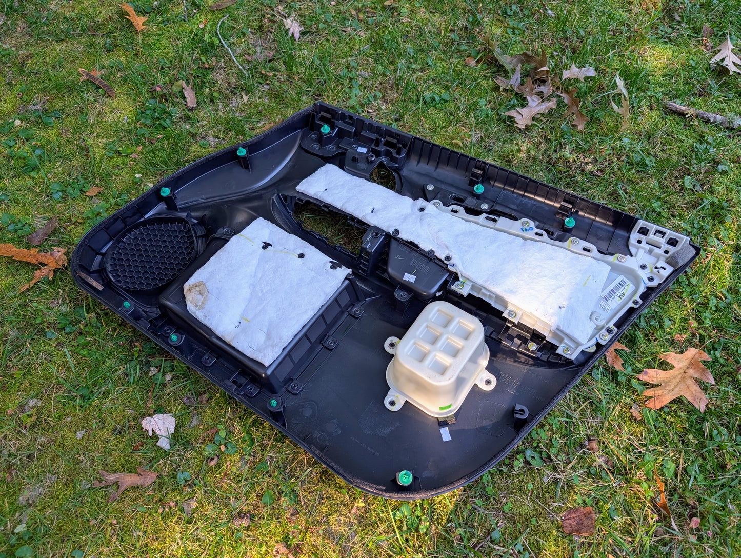 A Honda OEM Front Driver Power Door Panel for 2015-2020 Fit LX trim is lying on green grass with scattered brown leaves.