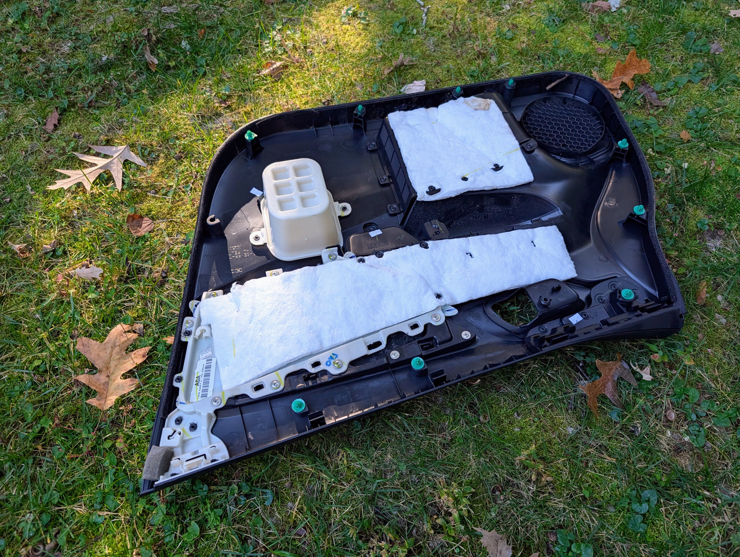 An OEM Honda Front Driver Power Door Panel for 2015-2020 Fit LX lies face down on green grass with brown leaves, sunlight highlighting its black surface and white insulation pads.