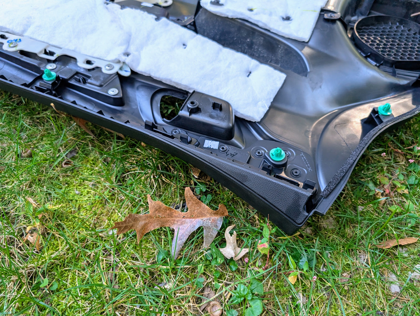 A black plastic OEM Honda front driver power door panel for 2015-2020 Fit LX lies on grass with insulation and green clips, near a brown oak leaf, apparently removed for repair or inspection.