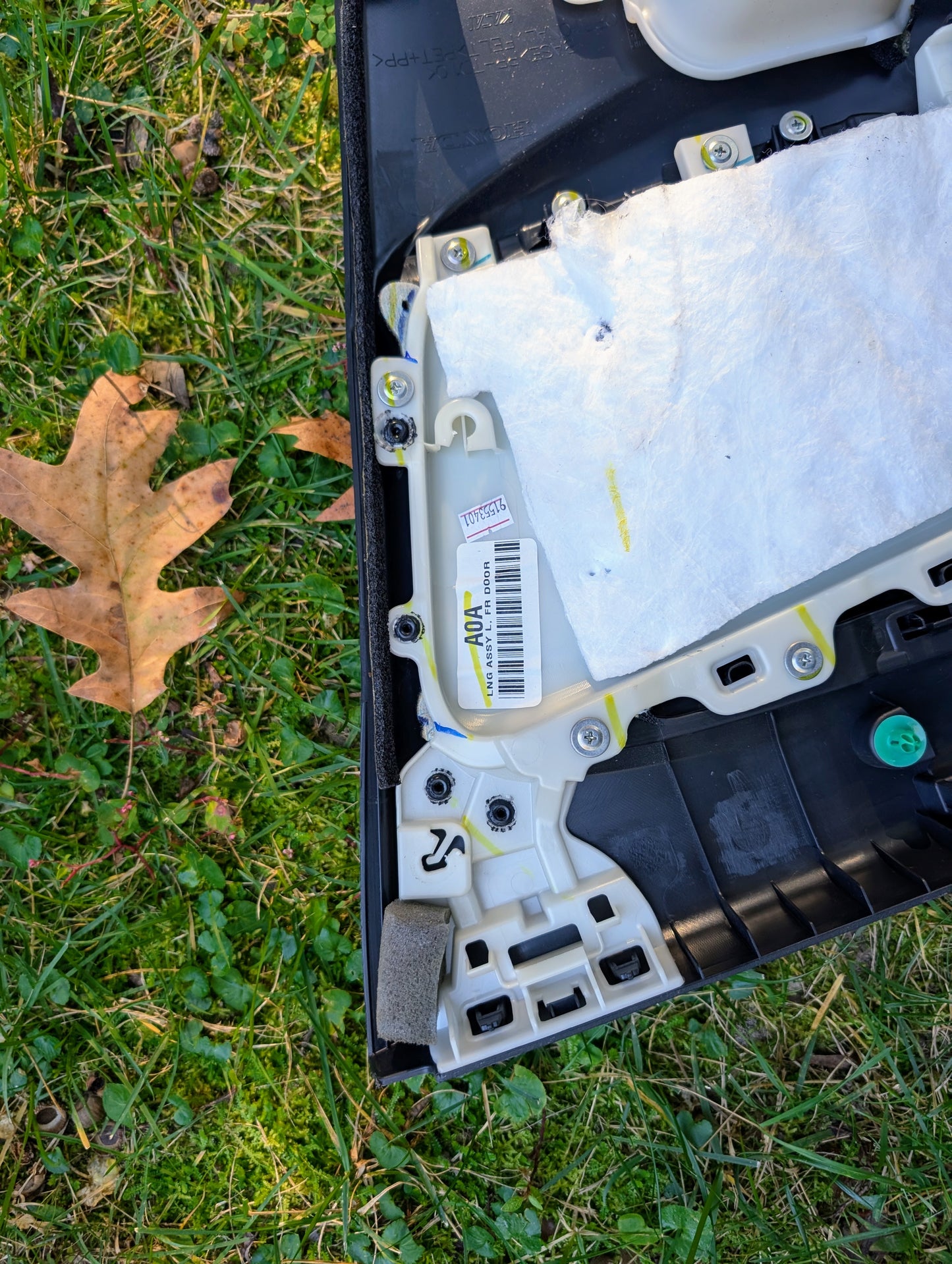 Close-up of a Honda OEM Front Driver Power Door Panel for 2015-2020 Fit LX on grass, displaying screws, a barcode, white padding, black plastic frame, and an oak leaf nearby.