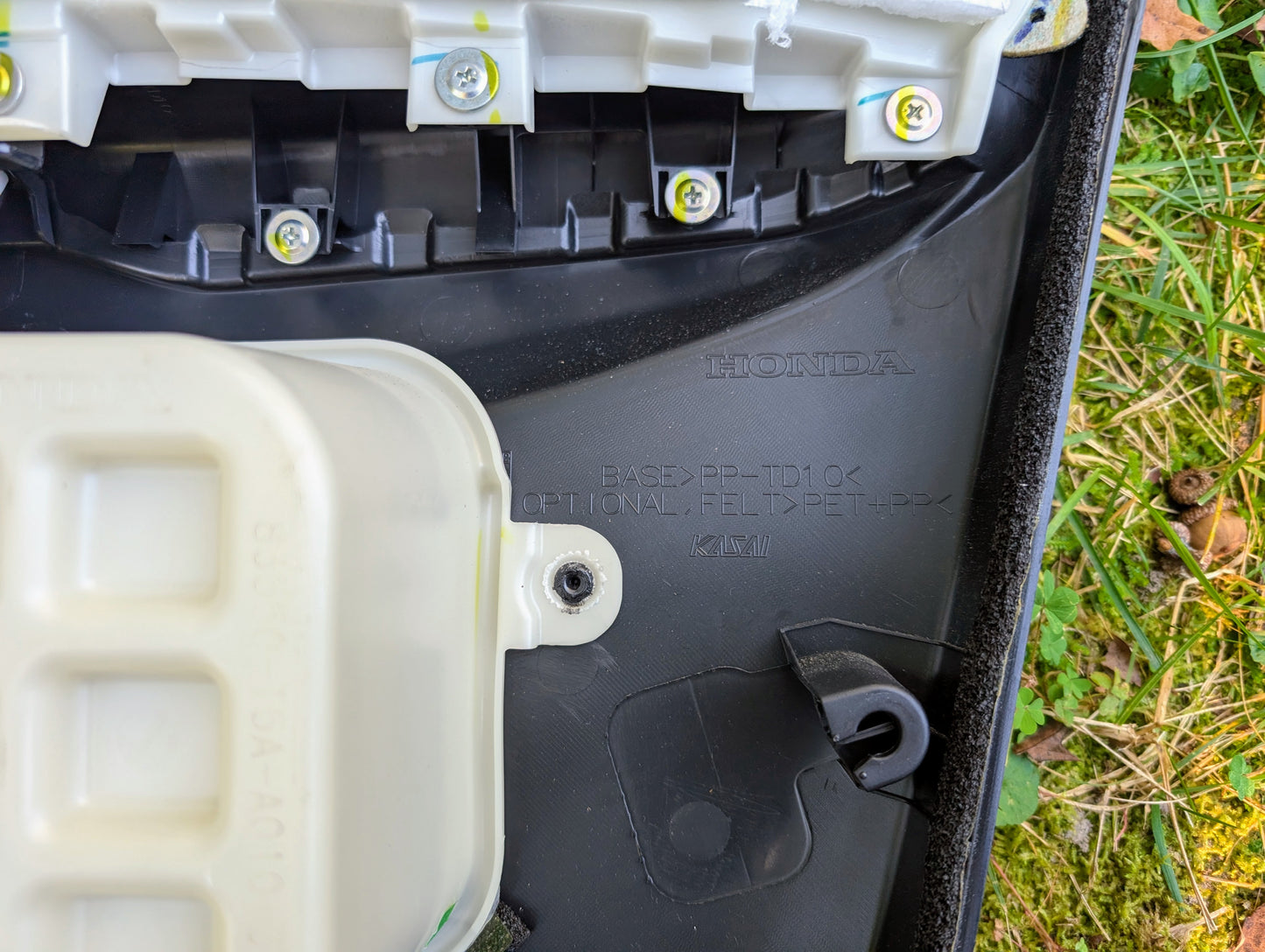 Close-up of an OEM Front Driver Power Door Panel for 2015-2020 Honda Fit LX by Honda, featuring black plastic, molded branding, a white component secured with screws, and grass visible in the background.