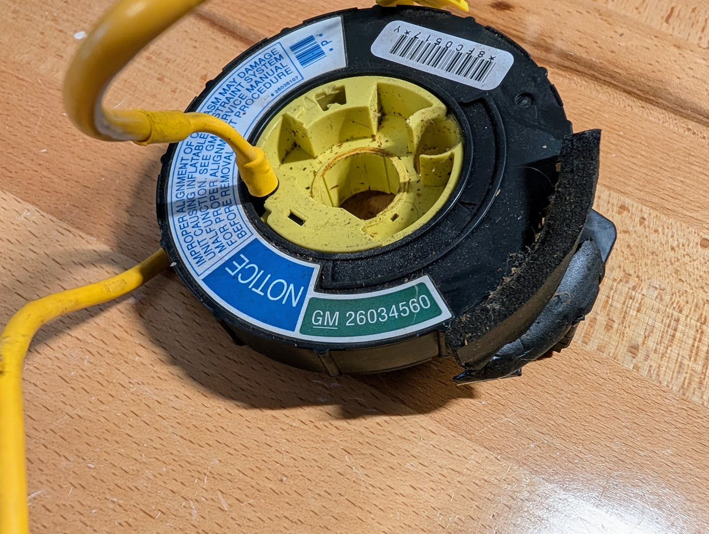 A close-up of a General Motors TESTED Steering Wheel SRS Air Bag Clock Spring OEM for 1995-1998 Chevy S10, C/K Truck, and more, with yellow wiring and part number GM 26034560, shown on a wooden surface.
