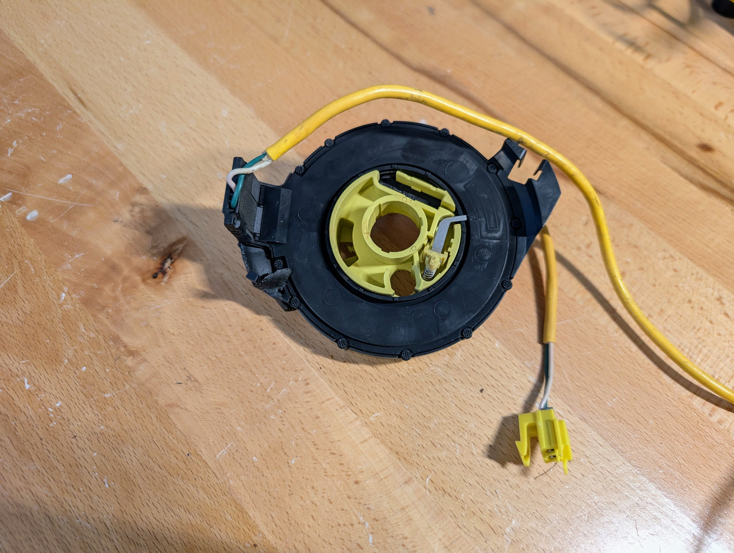 A tested OEM General Motors steering wheel SRS air bag clock spring for 1995-1998 Chevy S10 and C/K Truck by GM is shown on a wooden surface, its black plastic casing partially broken to reveal yellow internal components.