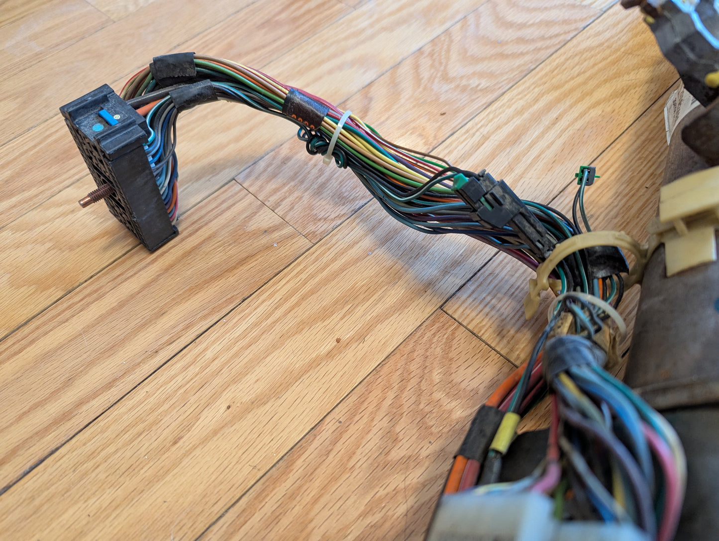 A bundle of multicolored wires, zip-tied together, connects to a black plug on a General Motors OEM Steering Column w/Tilt, Cruise Control, and Console Shift for 1998-2005 Chevy S10 Blazer and more, resting on a wooden floor.