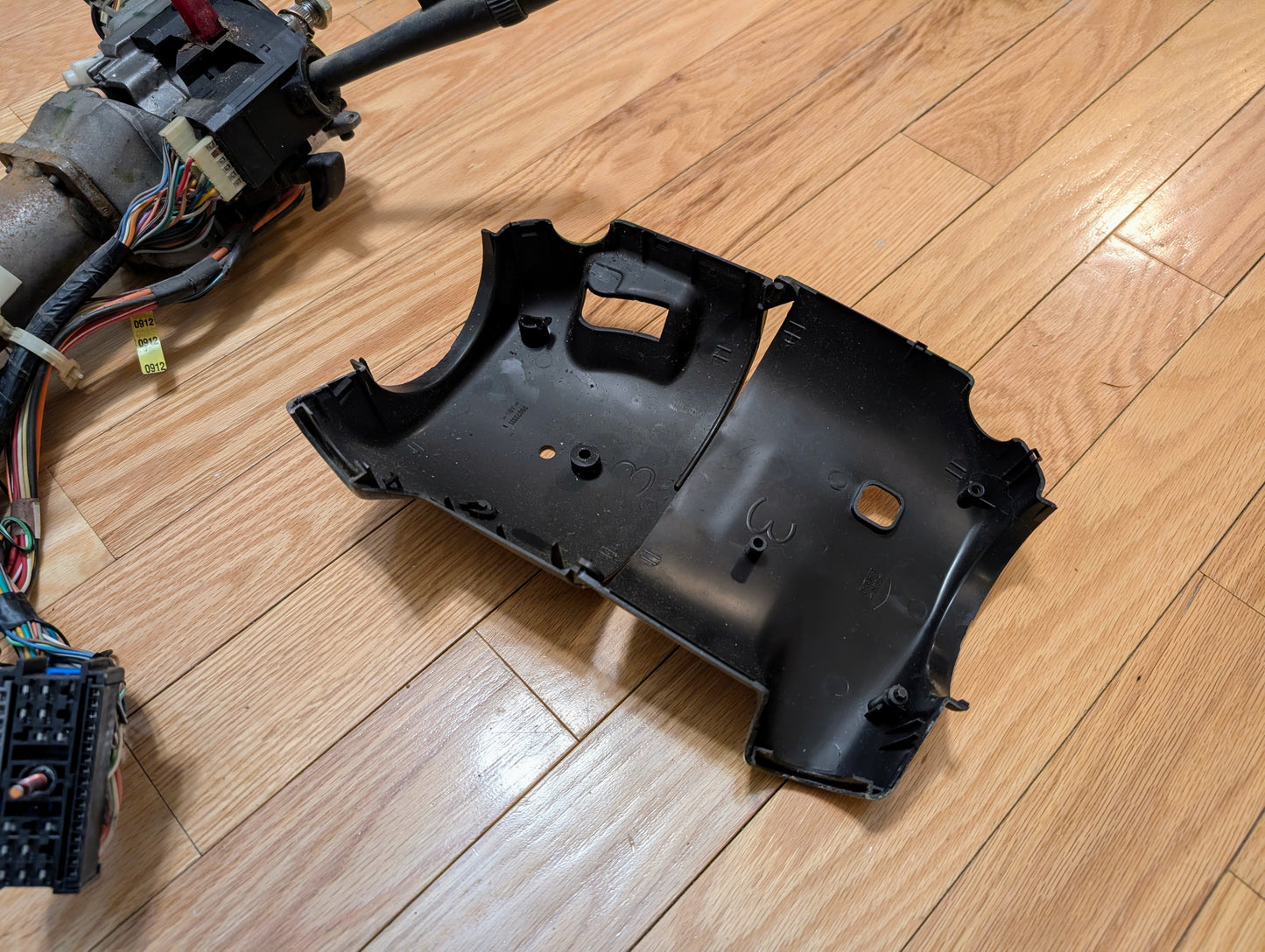 A black plastic steering column cover, in two pieces, rests on a light wood floor beside a General Motors OEM Steering Column w/Tilt, Cruise Control, and Console Shift for 1998-2005 Chevy S10 Blazer and more with exposed wiring and connectors.