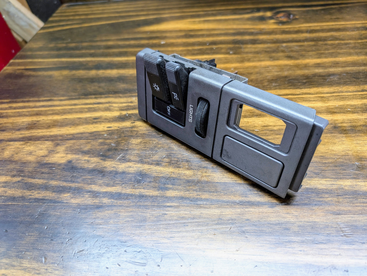 A close-up of a General Motors OEM Headlight Headlamp Switch for 1986-1994 Chevy S10, Blazer, GMC S15, and Jimmy shows the black dashboard panel with light control buttons and a LUMBAR button resting on a wood surface.