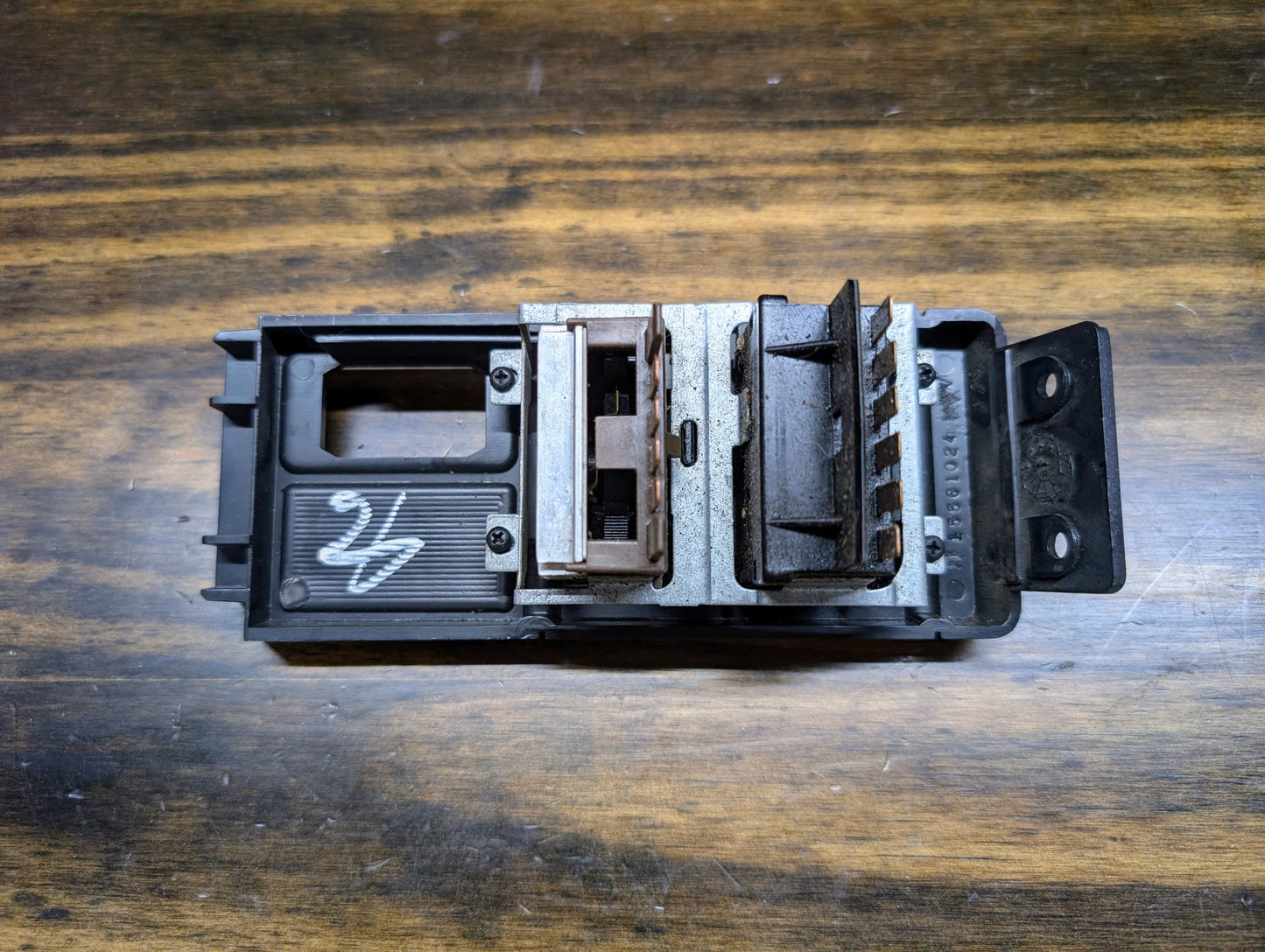 A close-up of a rectangular OEM Headlight Headlamp Switch by General Motors for 1986-1994 Chevy S10, Blazer, GMC S15, and Jimmy shows metal and plastic parts, slots, connectors, and a white marking on the left side on a dark wooden surface.