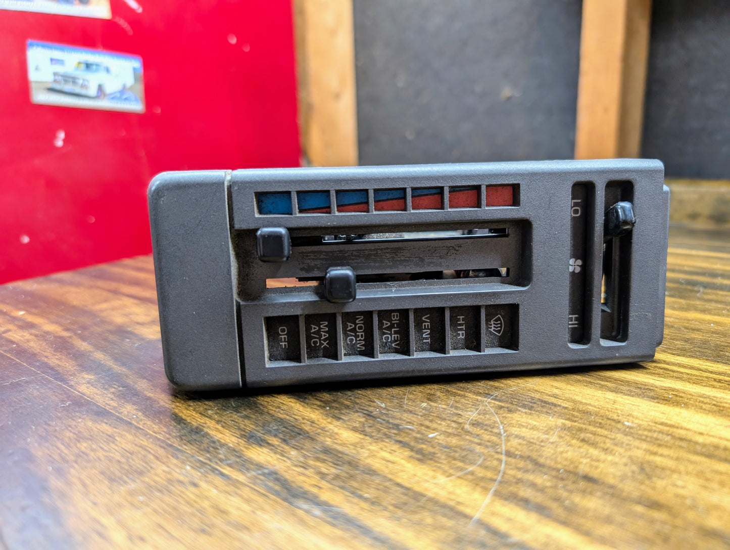 A close-up of a General Motors HVAC Heater Control Panel with A/C for 1986–1994 Chevrolet S10, Blazer, and more, featuring sliders for temperature, fan speed, and airflow direction, placed on wood with a red and gray background.