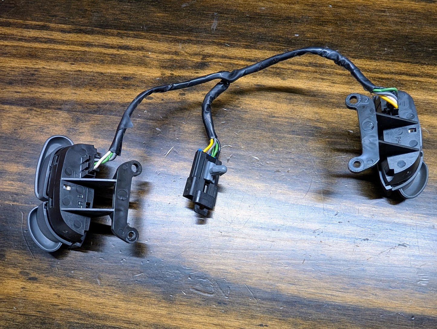Two black plastic electronic switches with wiring and a connector, resembling General Motors KIT Steering Wheel Radio Controls & Clock Spring for 1999 Chevy S10, Blazer, and more, are placed on a wooden surface.