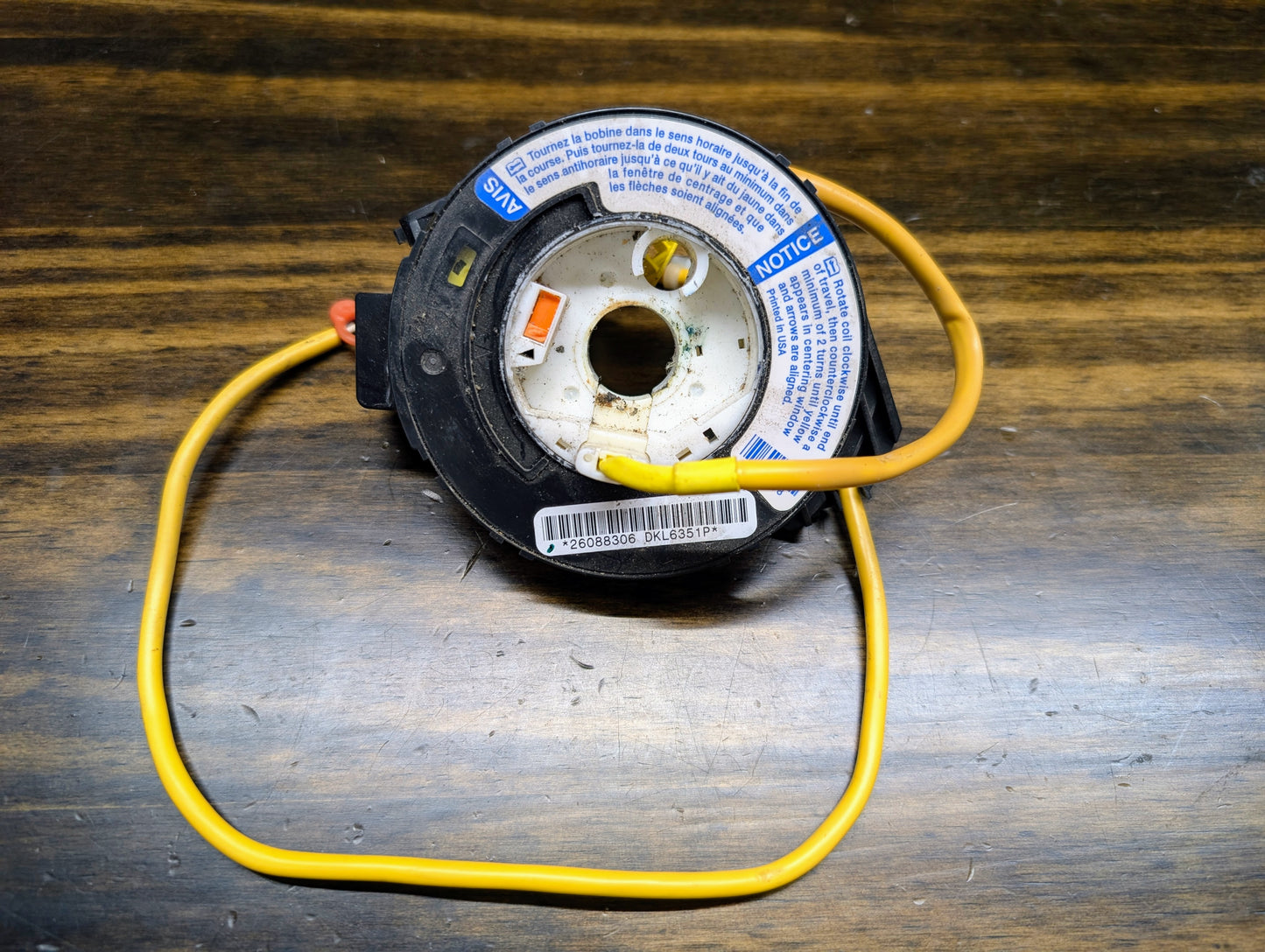 A close-up of a General Motors TESTED Steering Wheel Air Bag SRS Clock Spring OEM for 2000-2003 Chevy S10 Blazer with yellow wire on wood, showing multilingual warning labels and part numbers 26088306, 26087310, 26083473.