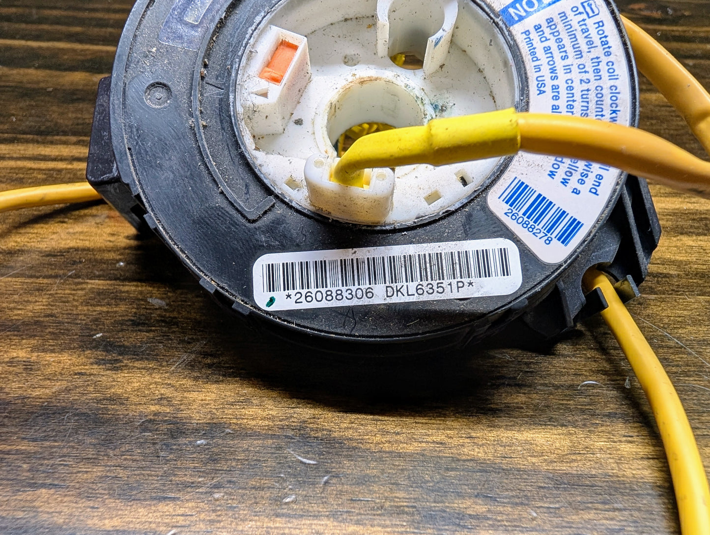 Close-up of a TESTED General Motors Steering Wheel Air Bag SRS Clock Spring OEM for 2000-2003 Chevy S10 Blazer, black and circular with yellow wires and barcode (26088306 DKL6351P), on wood surface.