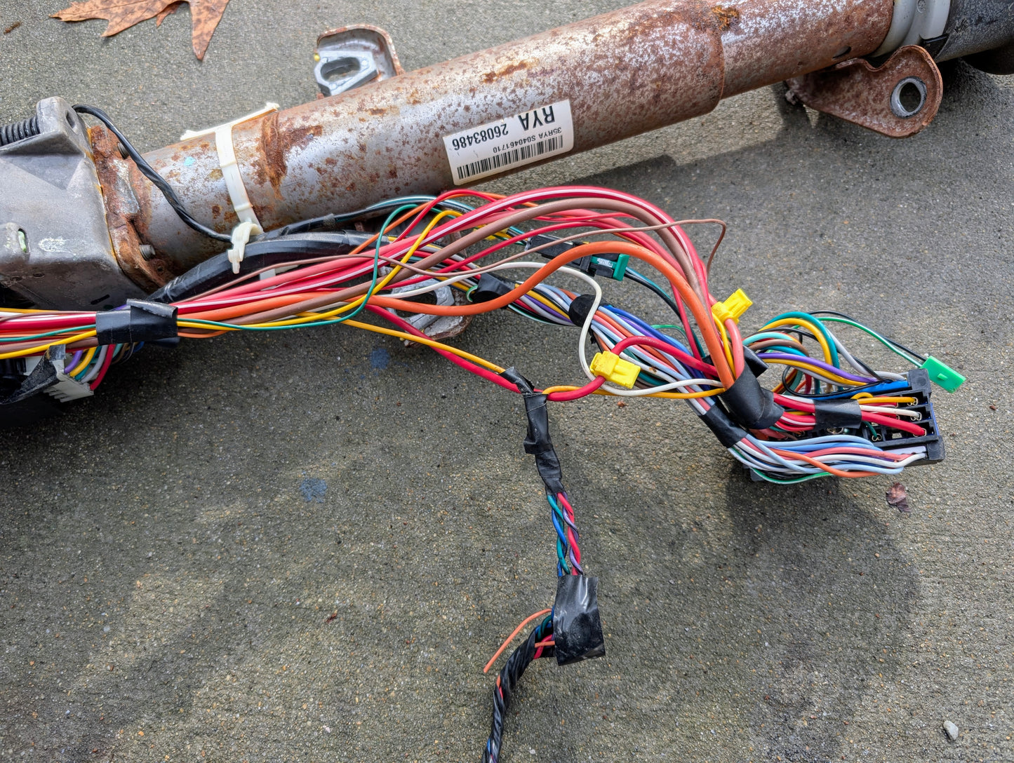 A bundle of multicolored wires with yellow connectors and black tape is attached to a rusted General Motors OEM Steering Column w/Tilt, Cruise Control, and Console Shift for 1998-2005 Chevy S10 Blazer, resting on concrete.