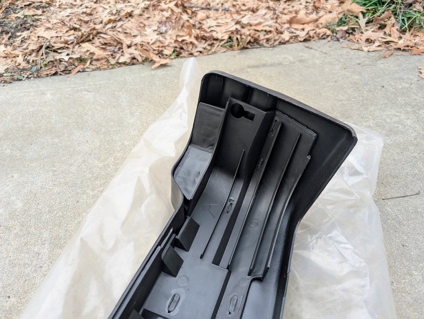 A General Motors NOS Rear Bumper Cover for 1991-1994 Oldsmobile Bravada lies on plastic sheeting over concrete, with dried leaves and grass in the background.