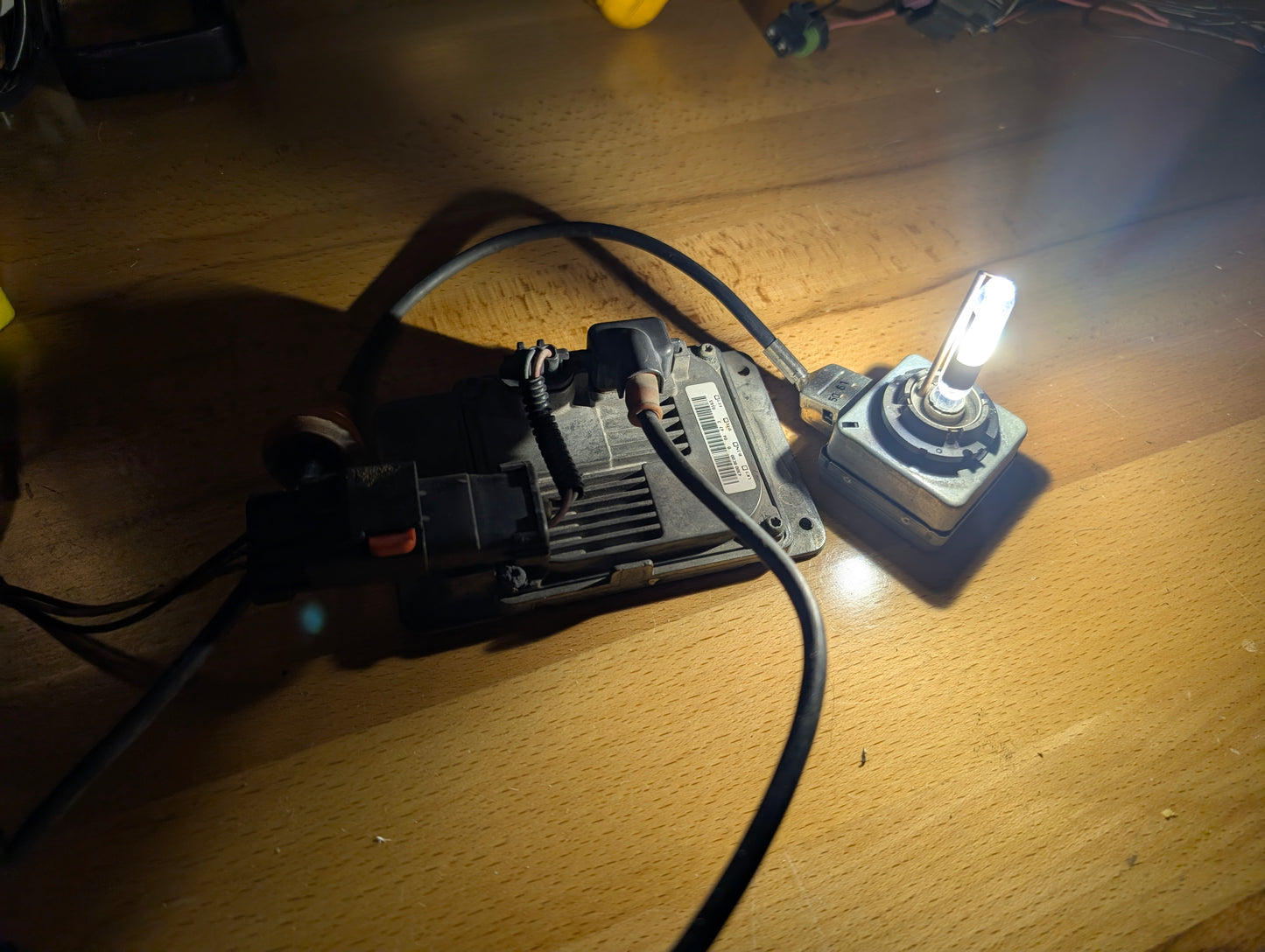 The General Motors OEM HID Headlamp Ballast 19121438, designed for 2002-2006 Cadillac Escalade, is shown powered and illuminating a wooden surface, casting bright light and shadows across the table.