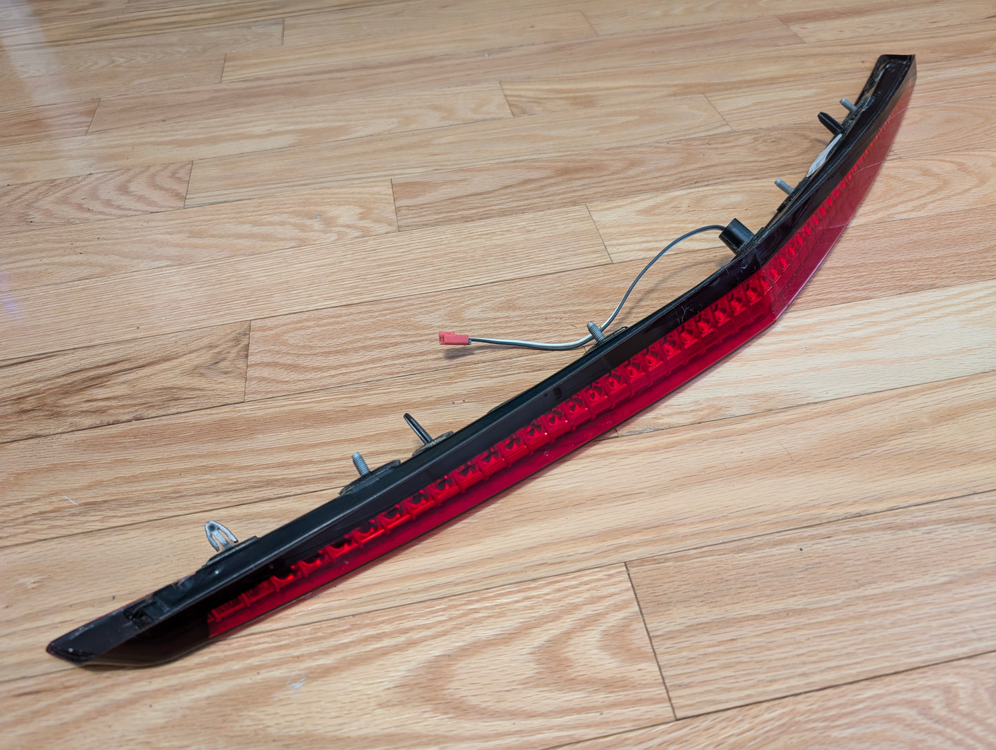 A General Motors OEM 2008-2013 Cadillac CTS Rear Stop Third Brake Light (CHMSL), with mounting clips and a wire connector, sits on a light wood floor—ideal for upgrading or replacing your Cadillac CTS trunk lamp.