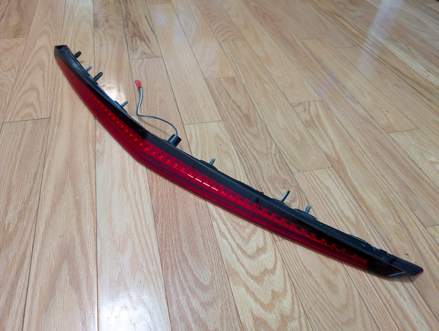 A General Motors OEM 2008-2013 Cadillac CTS Rear Stop Third Brake Light CHMSL with wiring connector is shown on a light wood floor—ideal for upgrading or replacing your CTS trunk lamp exterior parts.