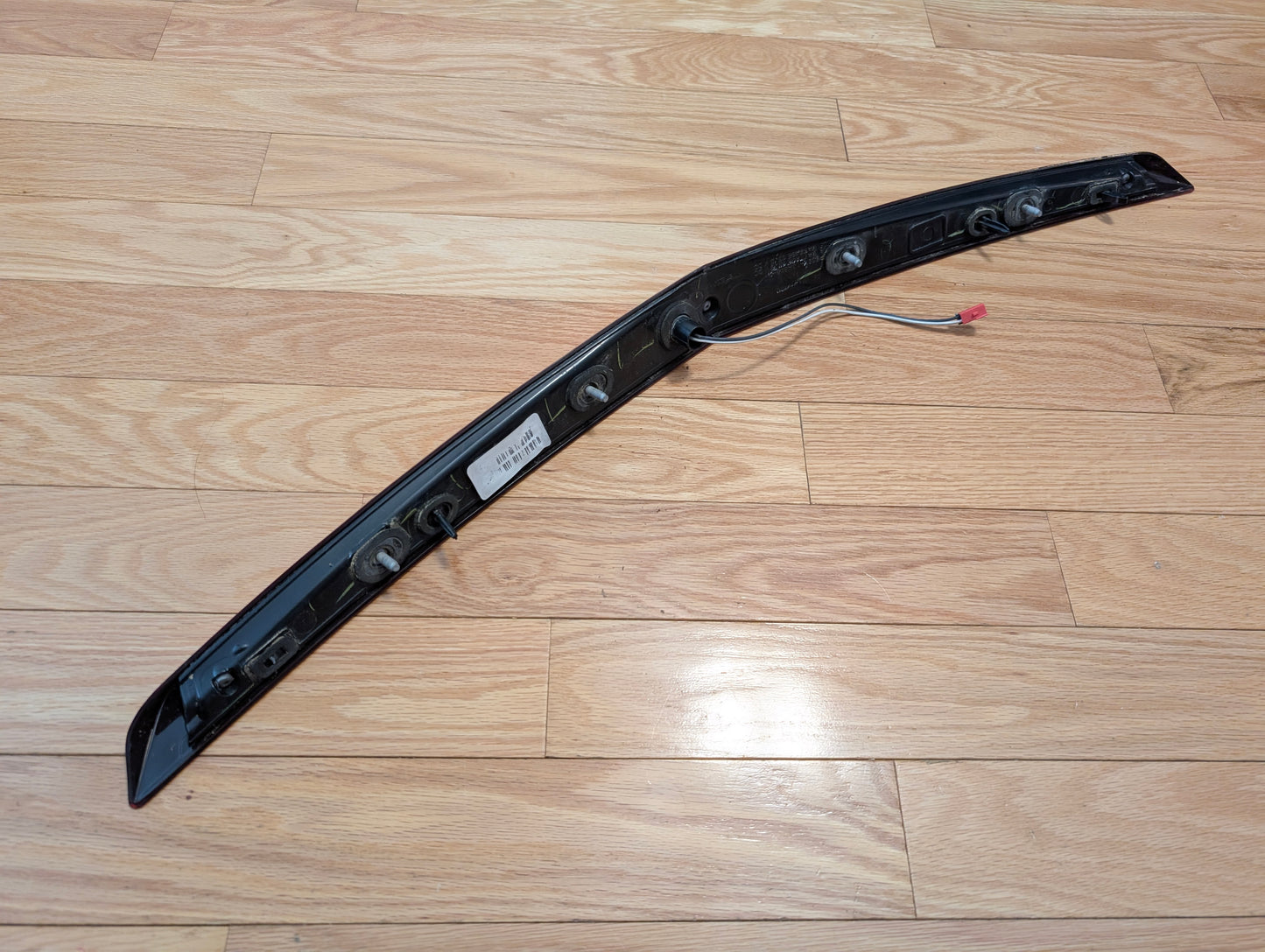 A General Motors OEM 2008-2013 Cadillac CTS Rear Stop Third Brake Light (CHMSL) with label and attached wiring sits on a light wood floor. The black plastic trunk lamp is long, narrow, slightly curved, and has multiple circular mounts for rear lighting.