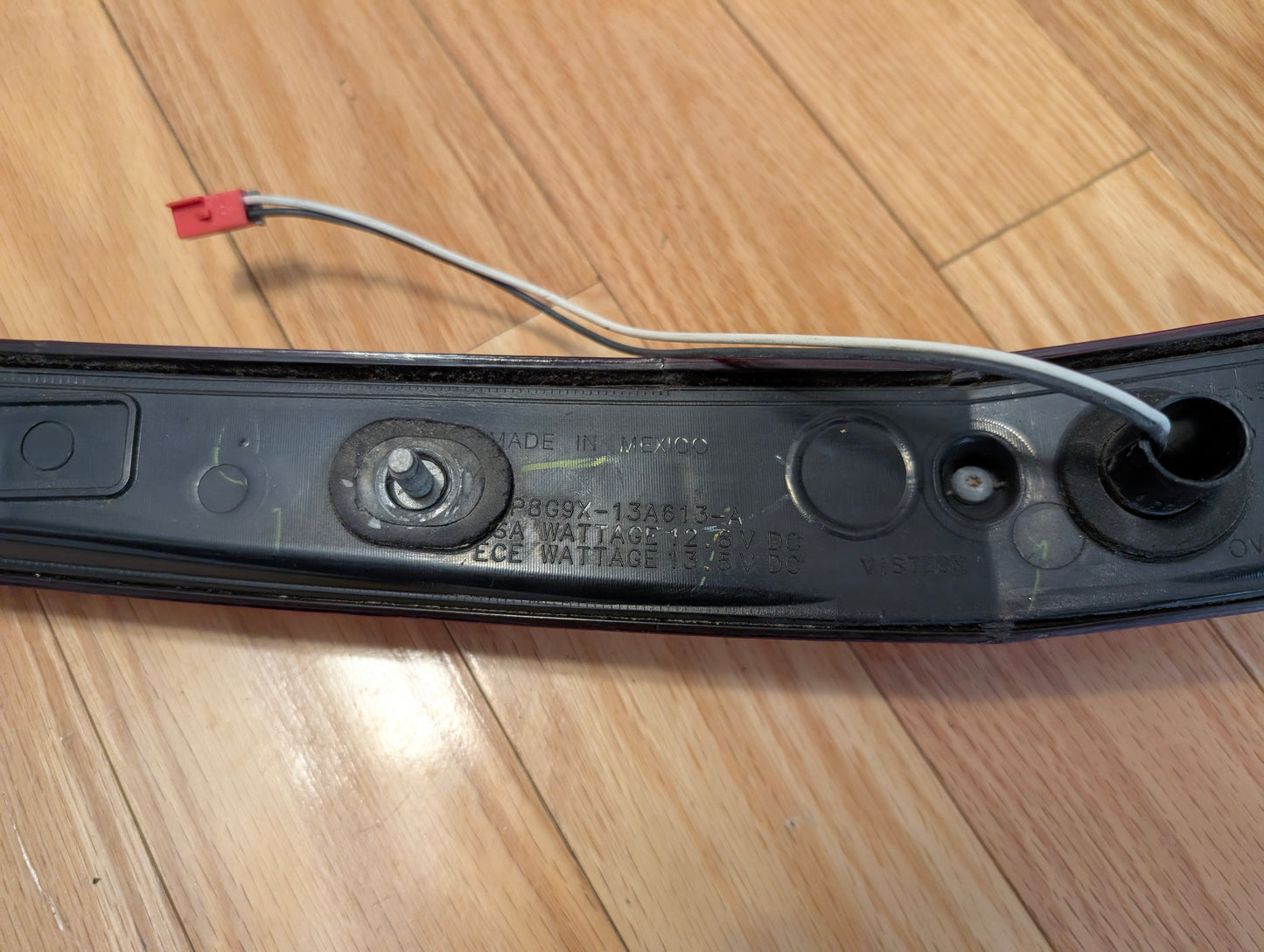 A close-up of a General Motors OEM 2008-2013 Cadillac CTS Rear Stop Third Brake Light (CHMSL) shows embossed specs, gray and white wire, and red connector, placed on a wooden floor.