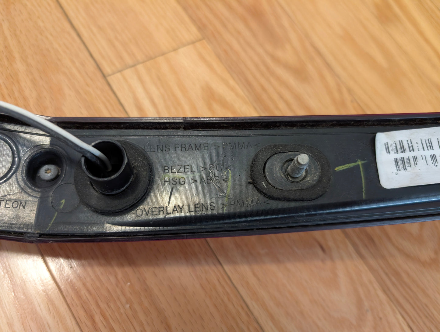 Close-up of a General Motors OEM 2008-2013 Cadillac CTS Rear Stop Third Brake Light CHMSL showing barcode, white wire, and engravings. Tested trunk lamp rests on wood floor—ideal for Cadillac CTS exterior replacement.