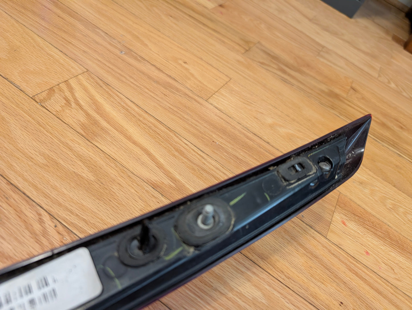 A close-up of a General Motors OEM 2008-2013 Cadillac CTS Rear Stop Third Brake Light (CHMSL) with bolts and connector, shown on a light wooden floor. Part of a barcode sticker is visible on the left side of this trunk lamp.