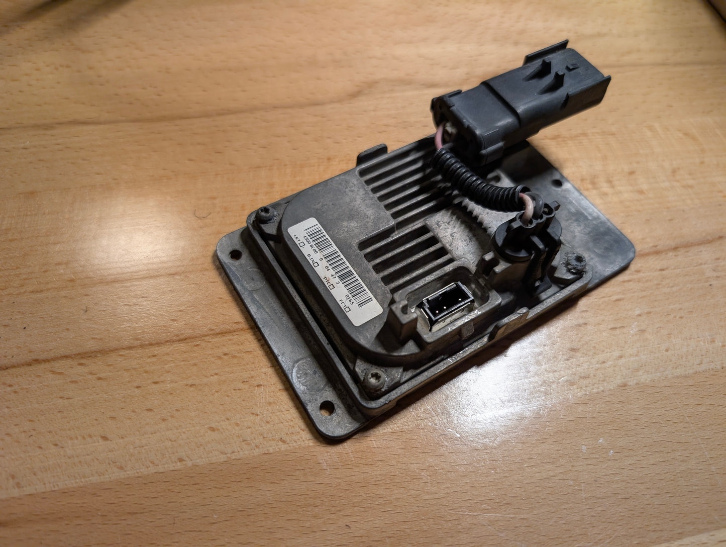 A close-up of a General Motors OEM HID Headlamp Ballast (part 19121438) for 2002-2006 Cadillac Escalade, showing its barcode label, connector port, and wiring on a light wooden surface—perfect for Escalade models.