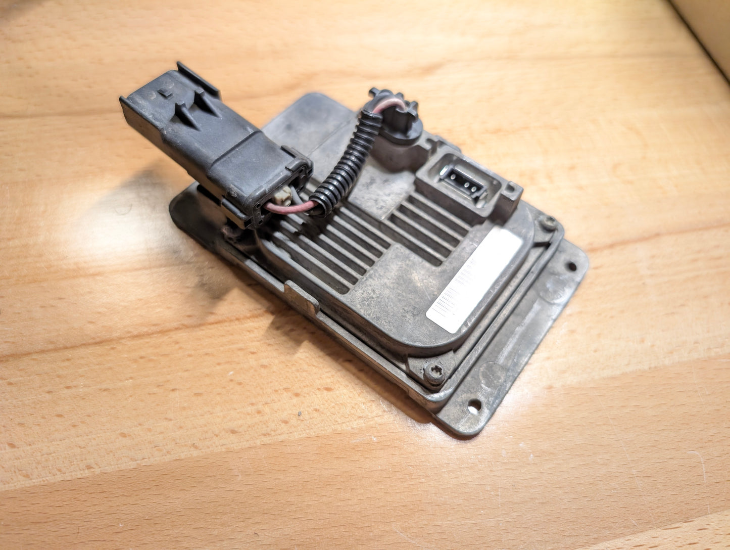 A close-up of the General Motors OEM HID Headlamp Ballast (19121438) for 2002-2006 Cadillac Escalade, featuring connectors and wires on a light wood surface—an essential rectangular module with black plastic connector.