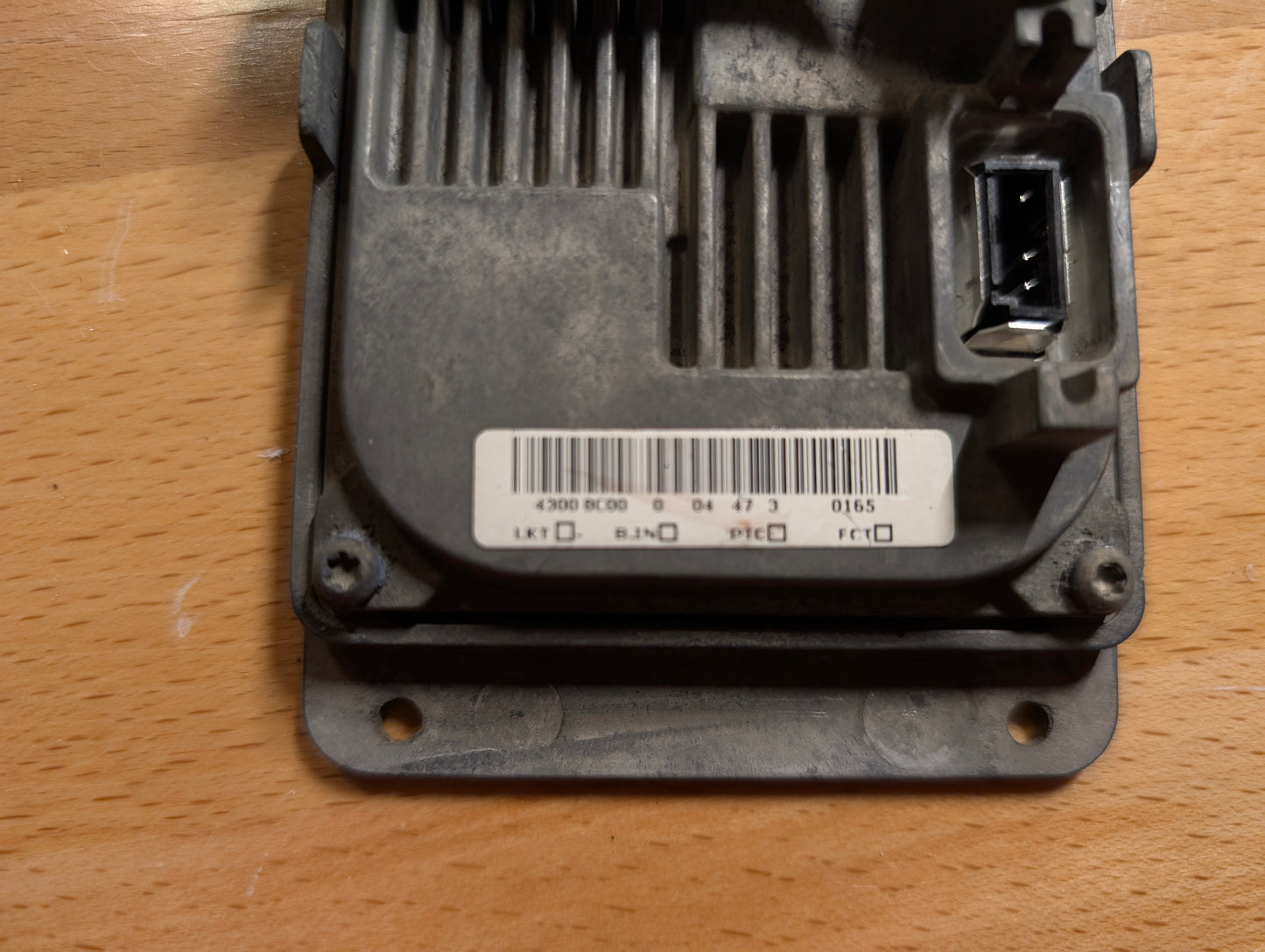 A dusty General Motors OEM HID Headlamp Ballast (19121438) for 2002-2006 Cadillac Escalade on a wooden surface, showing a barcode label and connector port on the right.