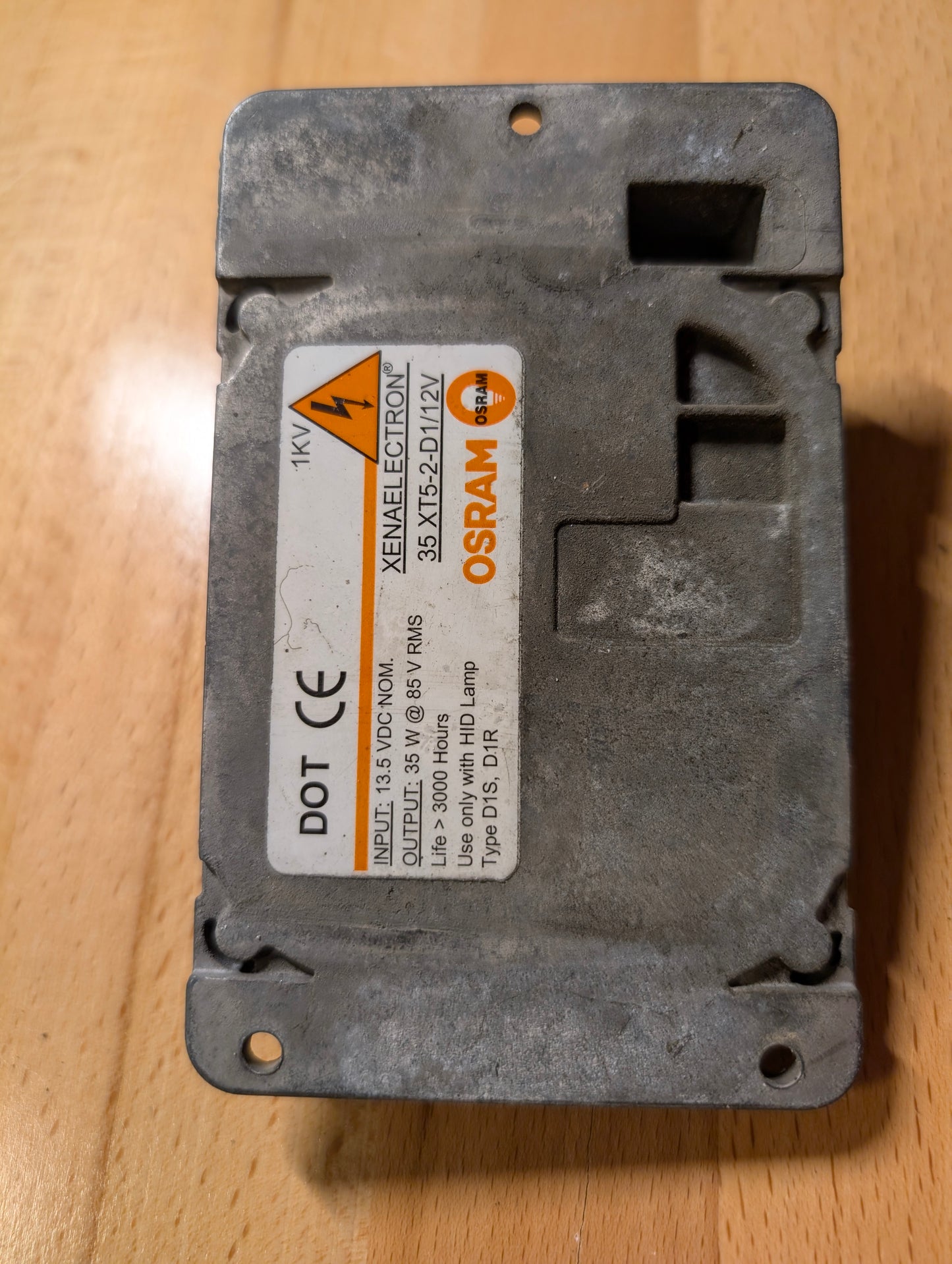A metallic General Motors OEM HID Headlamp Ballast (19121438) for 2002-2006 Cadillac Escalade with a worn label showing voltage, warnings, and certifications, resting on a wooden surface—fits many GM Full Size Trucks.