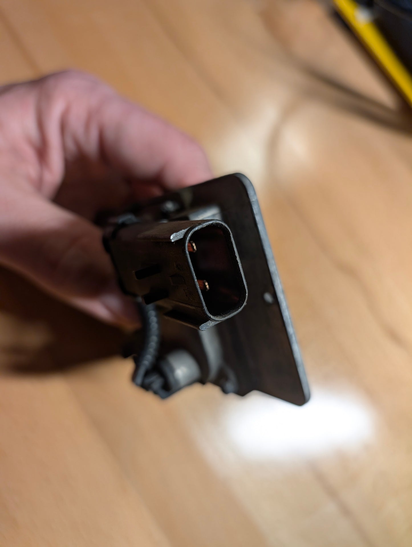 A close-up of a hand holding a General Motors OEM HID Headlamp Ballast (19121438) with metal mounting plate for 2002-2006 Cadillac Escalade, shown on a wooden surface.