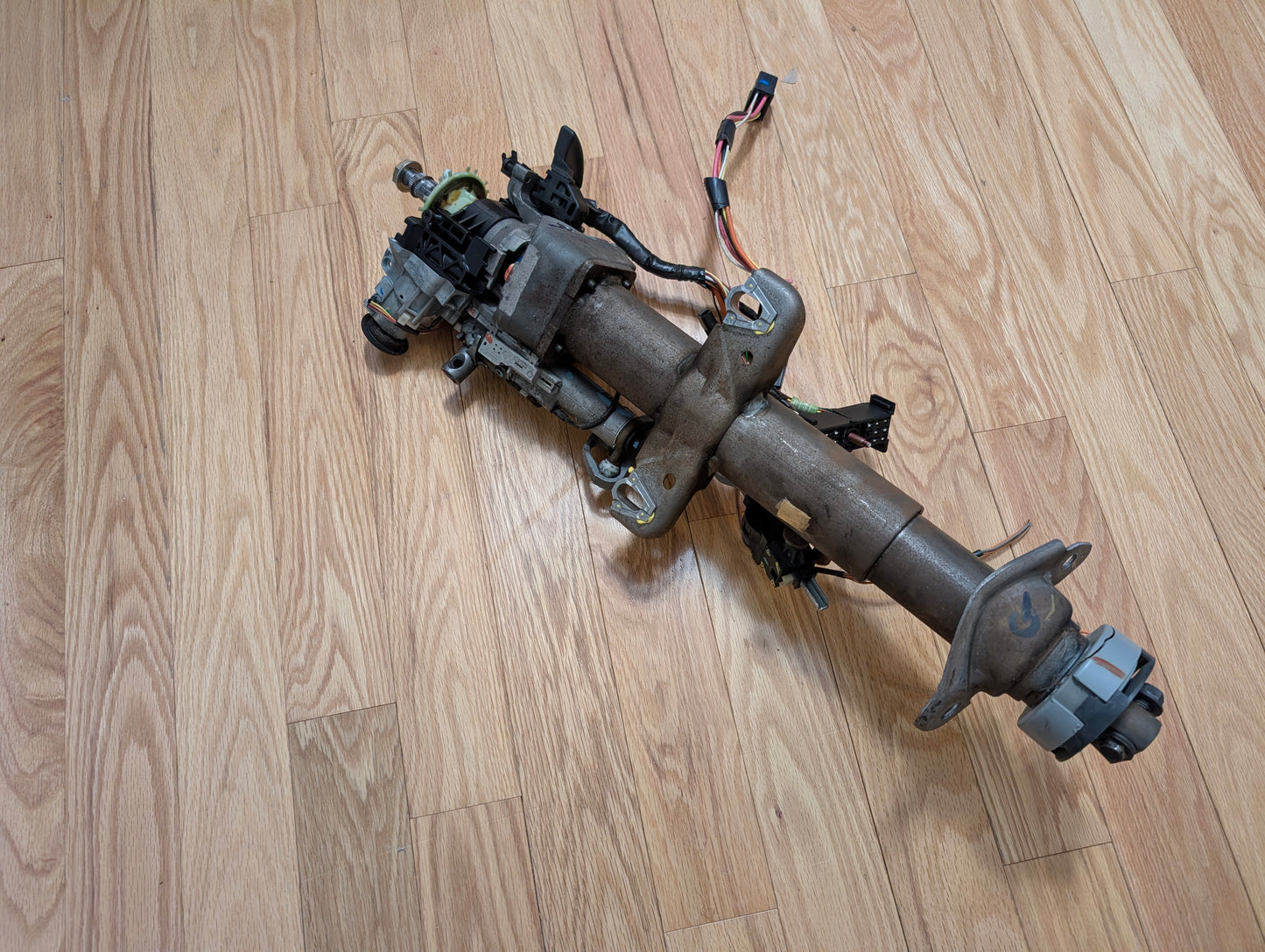 A General Motors OEM Steering Column w/KEY for 2000-2002 GM Full Size Trucks, designed for automatic transmissions and including wiring and a column shifter, is resting on a light wood floor.
