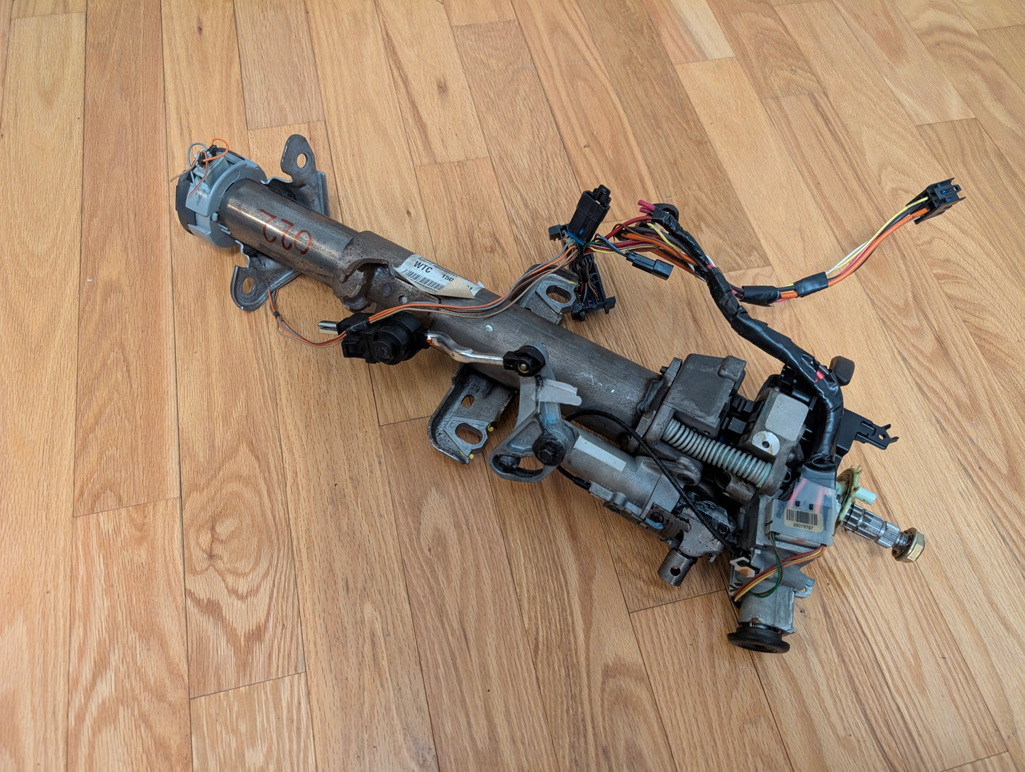An OEM Steering Column w/KEY for 2000-2002 GM Full Size Trucks by General Motors, featuring metal parts, brackets, attached wires, connectors, and a column shifter for automatic transmission, is placed on a wooden floor.