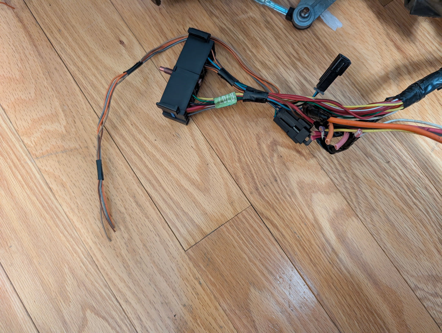 An OEM Steering Column w/KEY by General Motors for 2000-2002 GM Full Size Trucks lies on a wooden floor, featuring multicolored wires, connectors, and a green fuse. Some wires are detached or stripped with various plastic connectors visible.