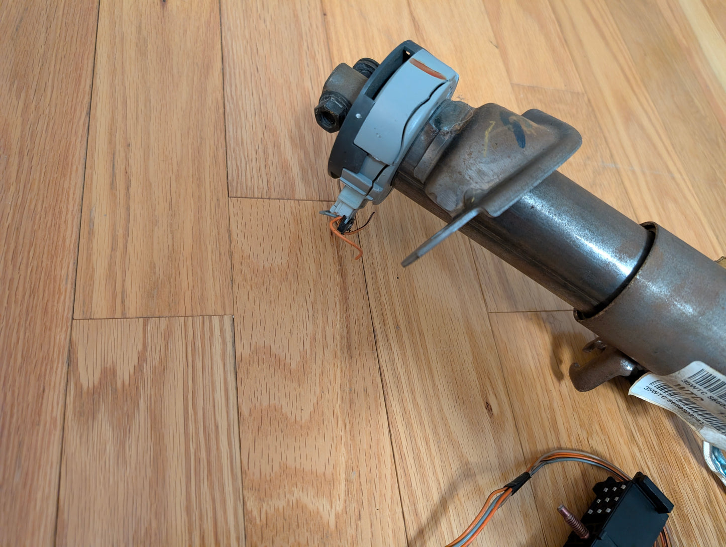 A close-up of an OEM Steering Column w/KEY for 2000-2002 GM Full Size Trucks by General Motors, showing electrical wires and a connector attached to the metallic cylindrical part, resting on a wooden floor.
