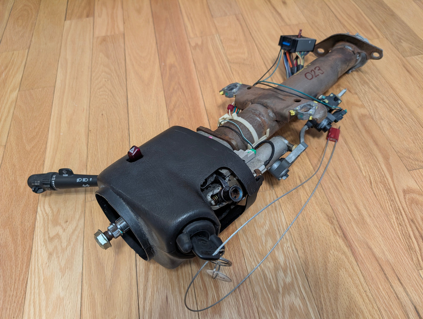A General Motors OEM Steering Column w/Tilt, Cruise Control, and Column Shift for 1998-2005 Chevy S10 Blazer and more is shown on a wooden floor, featuring attached wiring, control lever, connectors, and a black plastic cover.
