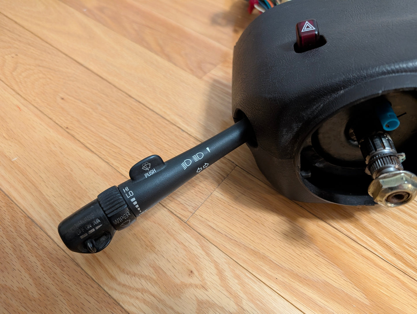 A close-up of a General Motors OEM Steering Column w/Tilt, Cruise Control, and Column Shift for 1998-2005 Chevy S10 Blazer shows its levers and wiring in a black housing resting on a wooden floor.