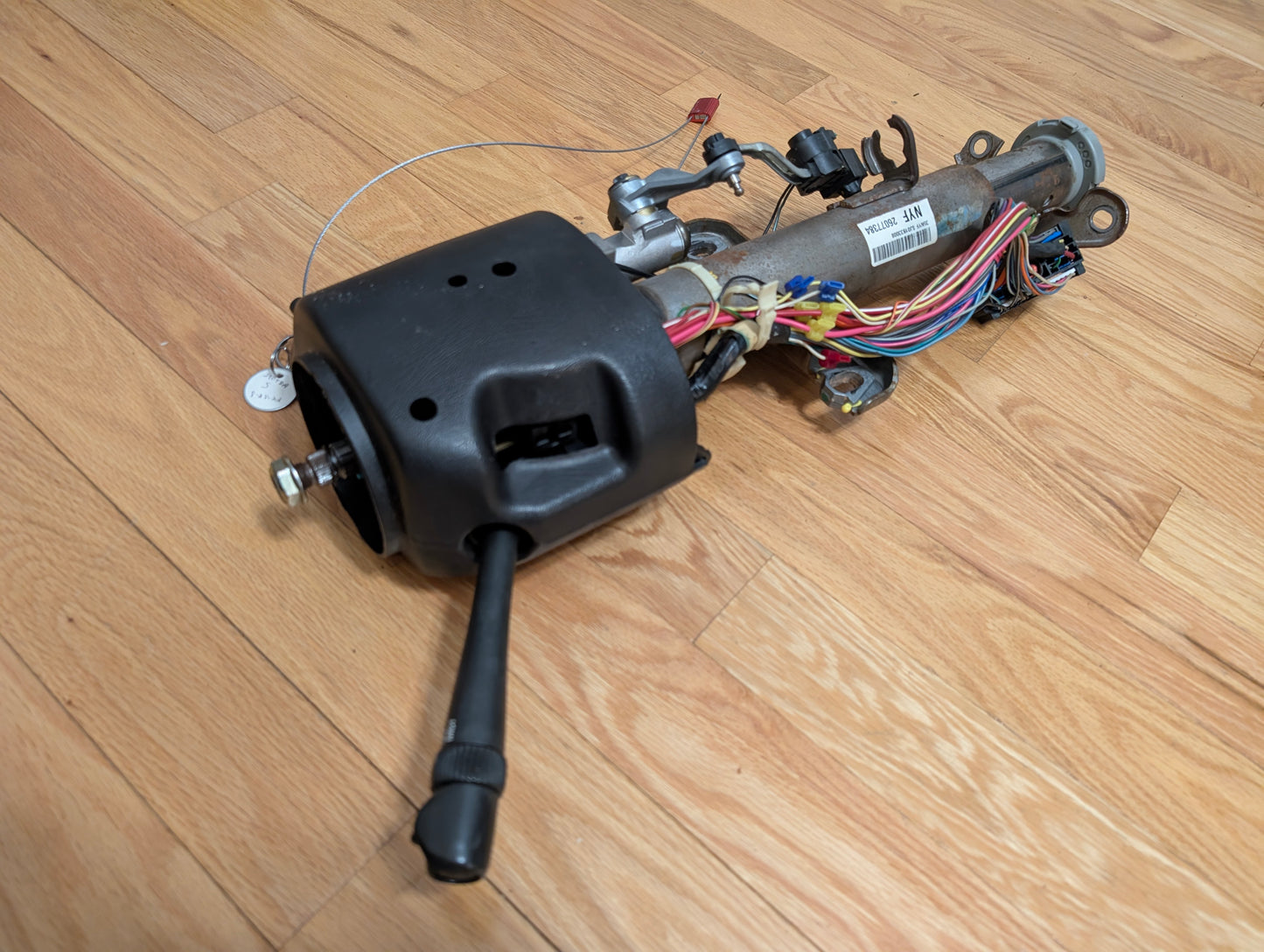 An OEM General Motors Steering Column w/Tilt, Cruise Control, and Column Shift for 1998-2005 Chevy S10/Blazer lies partially disassembled with wiring and lever on a wooden floor; the black plastic cover is partially attached.