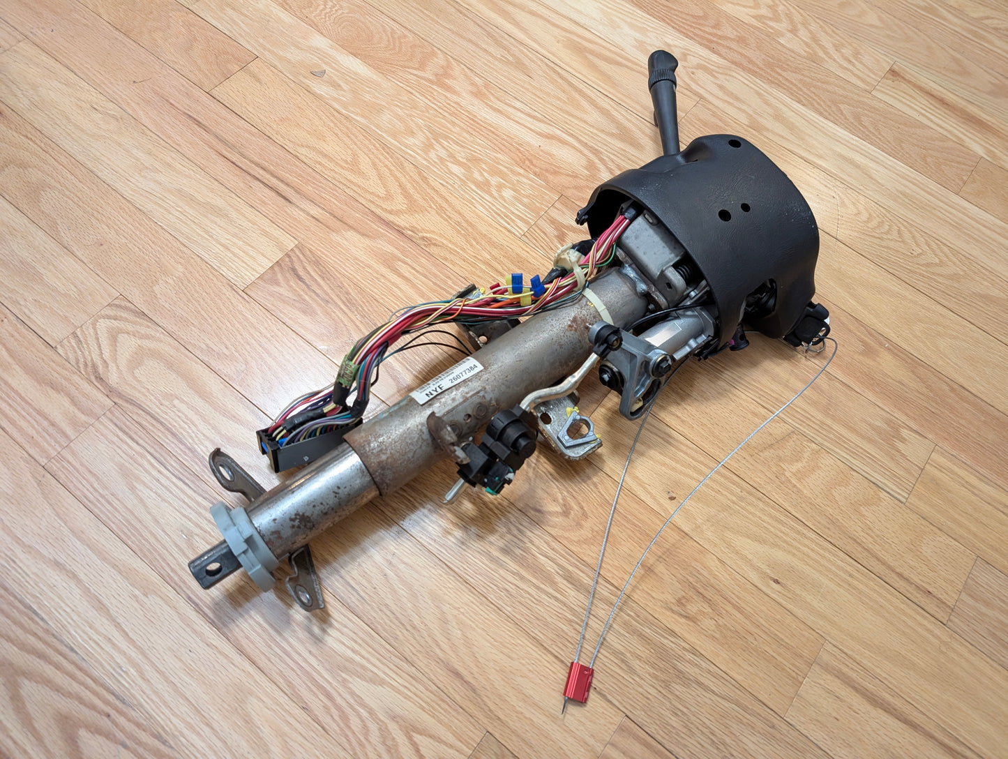 A General Motors OEM Steering Column w/Tilt, Cruise Control, and Column Shift for 1998-2005 Chevy S10 Blazer rests on a wooden floor, showing the metal shaft, wiring harness, and black plastic housing.