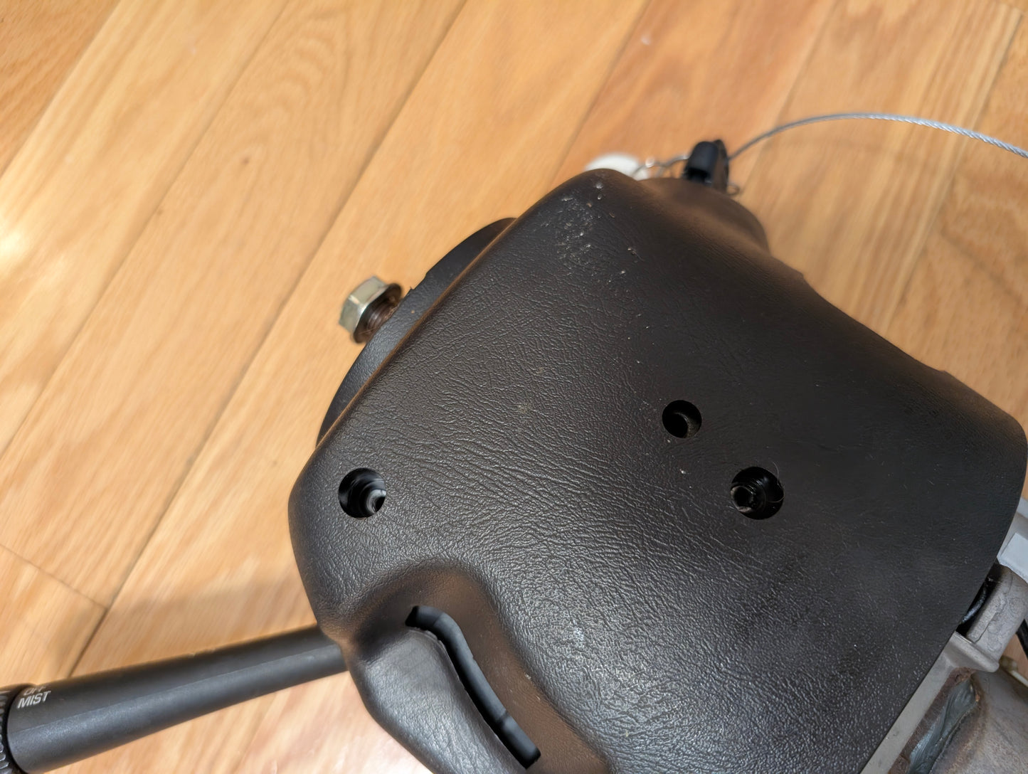 A close-up of a General Motors OEM Steering Column w/Tilt, Cruise Control, and Column Shift for 1998-2005 Chevy S10 Blazer shows two holes and a lever, with a cable and cruise control knob in the background on a wooden floor.