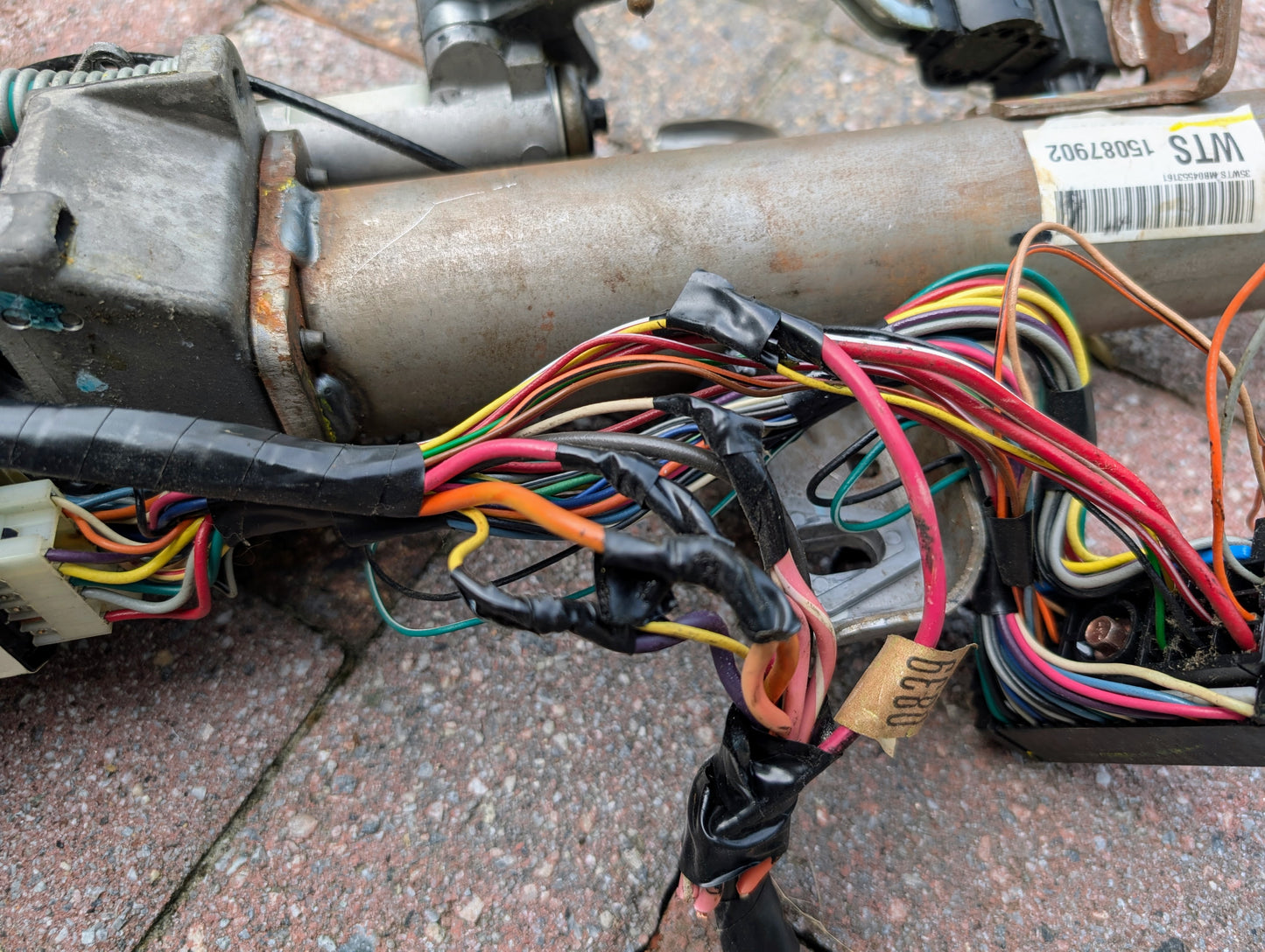 Close-up of a bundle of multicolored wires wrapped in black tape, attached to an OEM Steering Column for 2000-2002 GM Full Size Trucks by General Motors, resting on a textured brick surface.