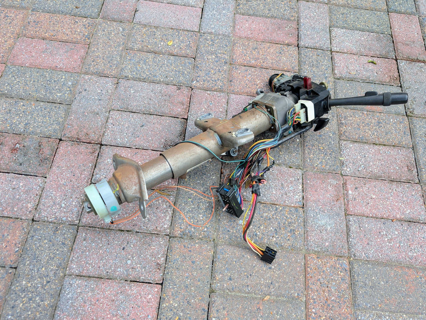 A General Motors OEM Steering Column for 2000-2002 GM Full Size Trucks, featuring exposed wiring and connectors, is displayed on a surface of red and gray bricks.