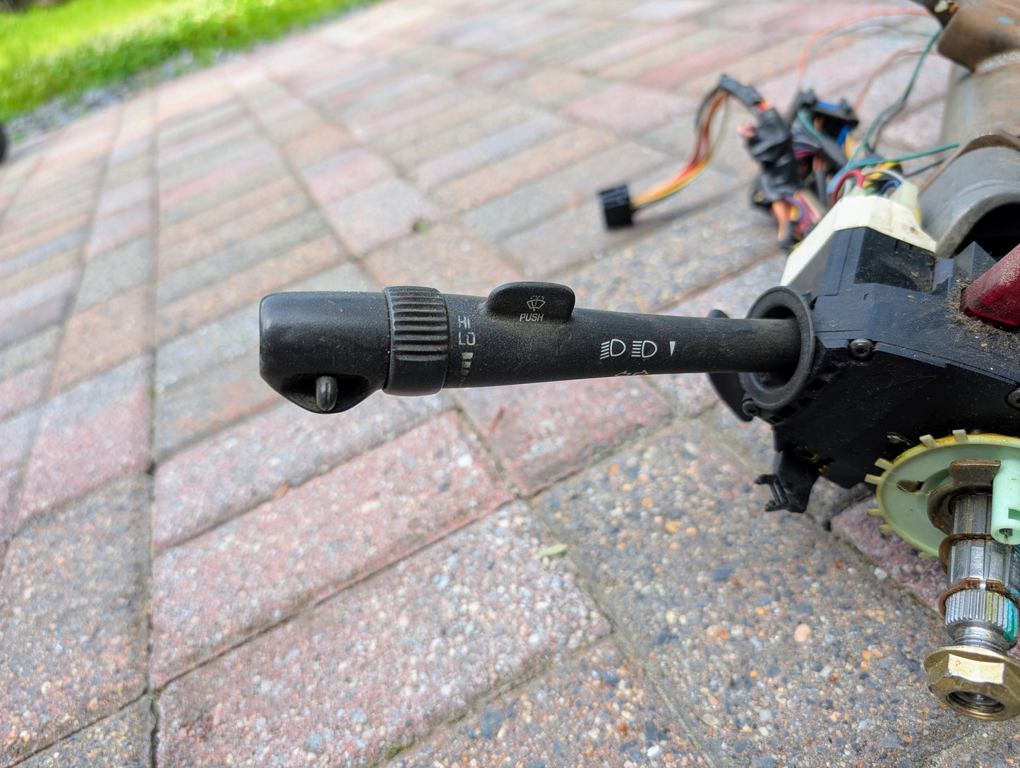 A close-up of a General Motors OEM Steering Column for 2000-2002 GM Full Size Trucks, featuring turn signal and wiper controls, is shown on a paved surface with visible wiring and mechanical parts.
