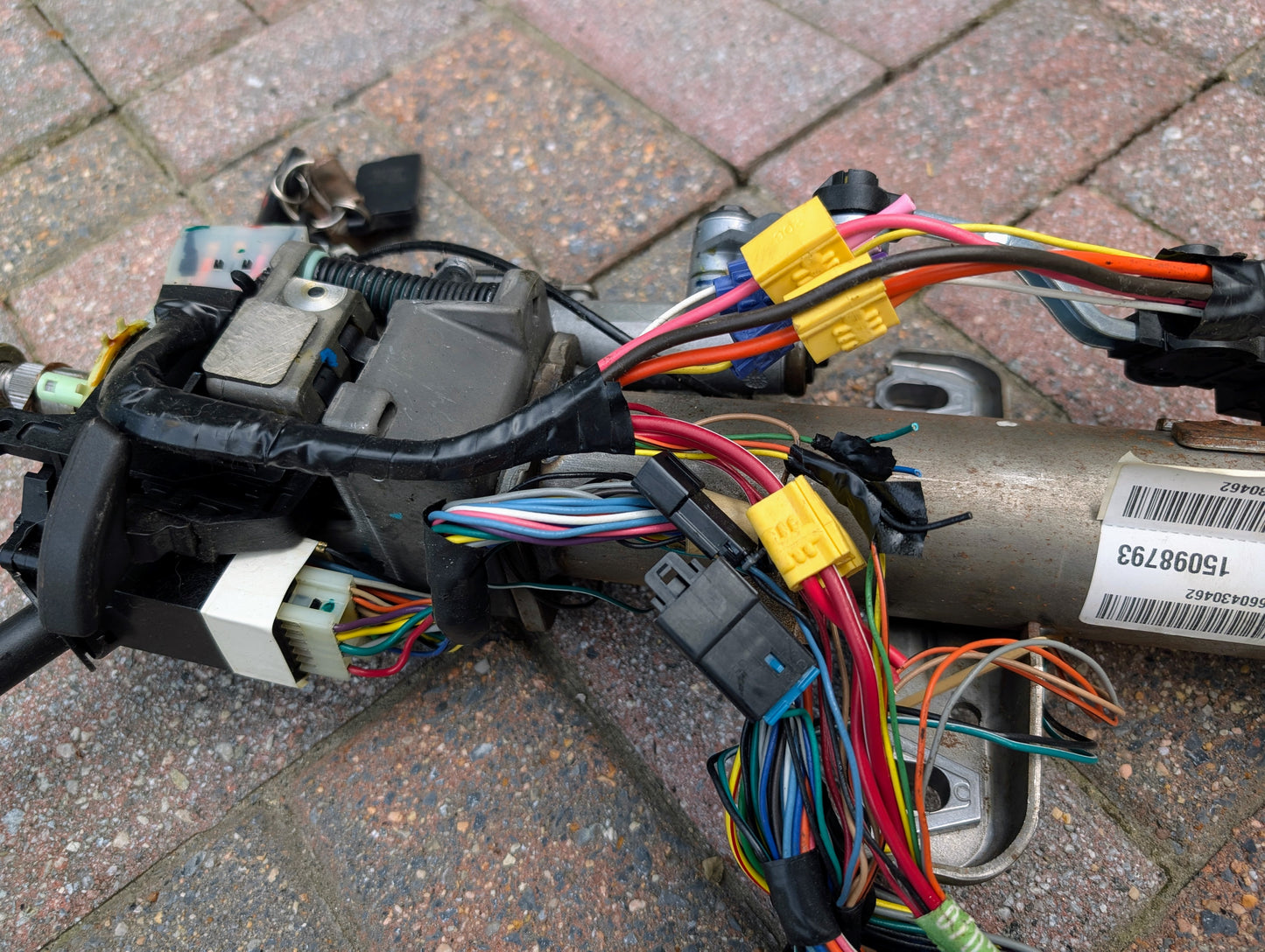 Close-up of a General Motors OEM Steering Column w/KEY for 2000-2002 GM Full Size Trucks, showing ignition switch assembly, colorful wires, connectors, car key, barcode label, and steering column components on brick pavement.