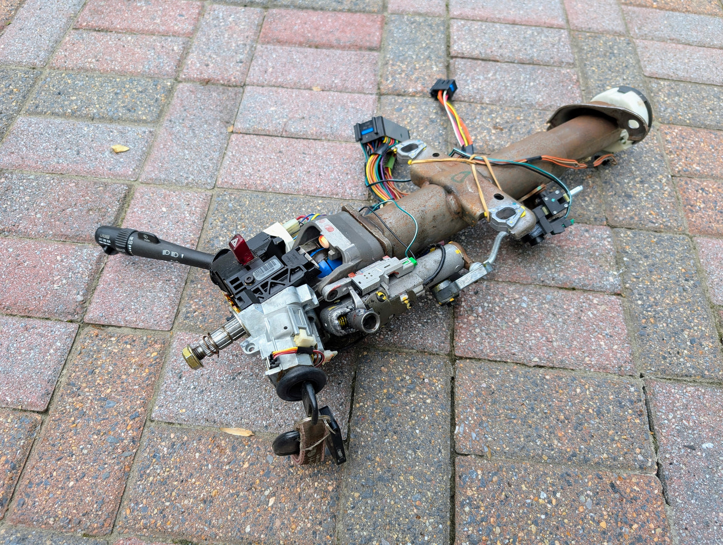 A General Motors OEM Steering Column w/KEY for 2000-2002 GM Full Size Trucks is lying on a brick-paved surface, complete with attached wiring and switches.