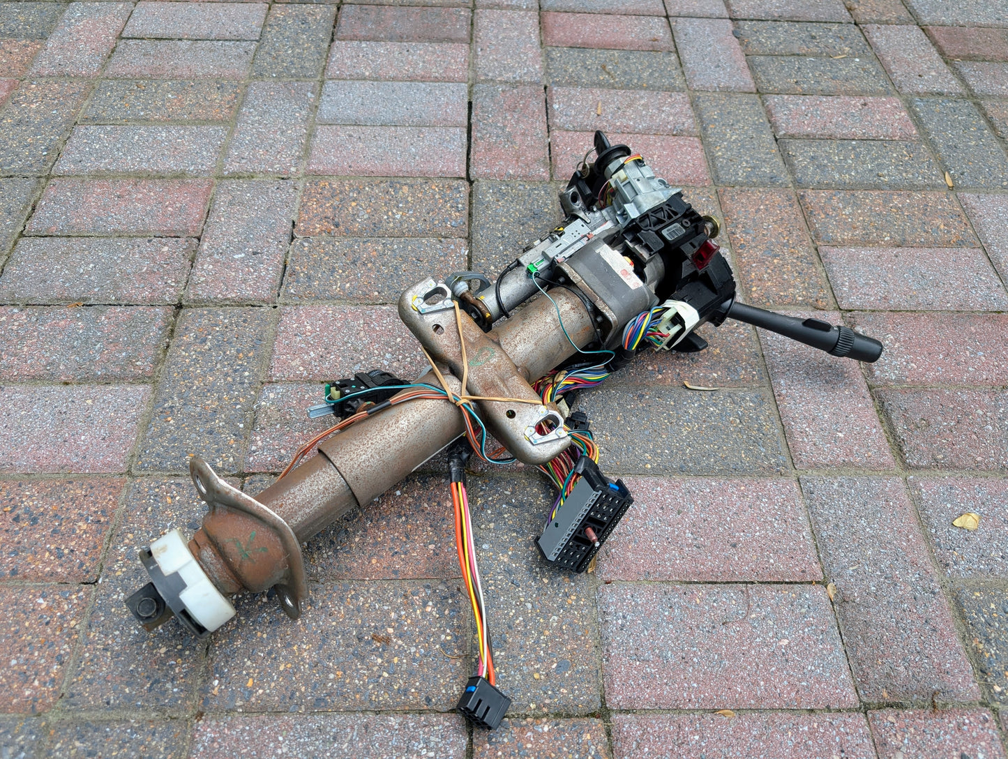 A rusty General Motors OEM Steering Column w/KEY for 2000-2002 GM Full Size Trucks, with exposed wires and connectors, rests on a patterned brick pavement.