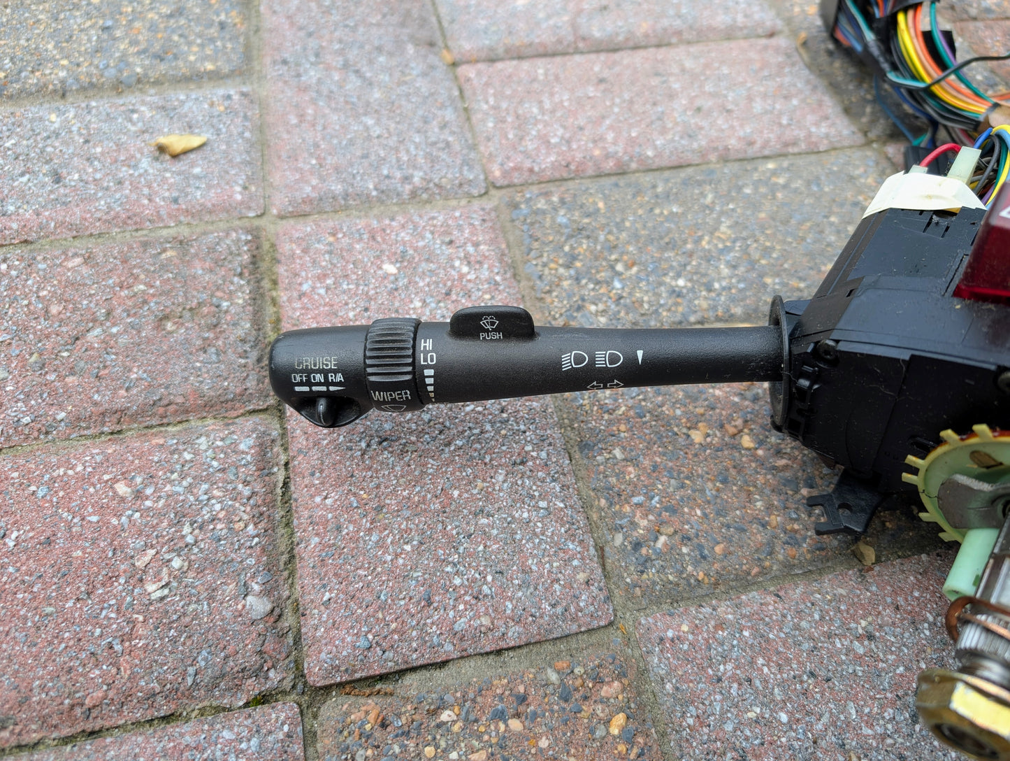 A close-up of a General Motors OEM Steering Column w/KEY for 2000-2002 GM Full Size Trucks, showing the multifunction switch with exposed wiring and parts, placed on stone pavement.