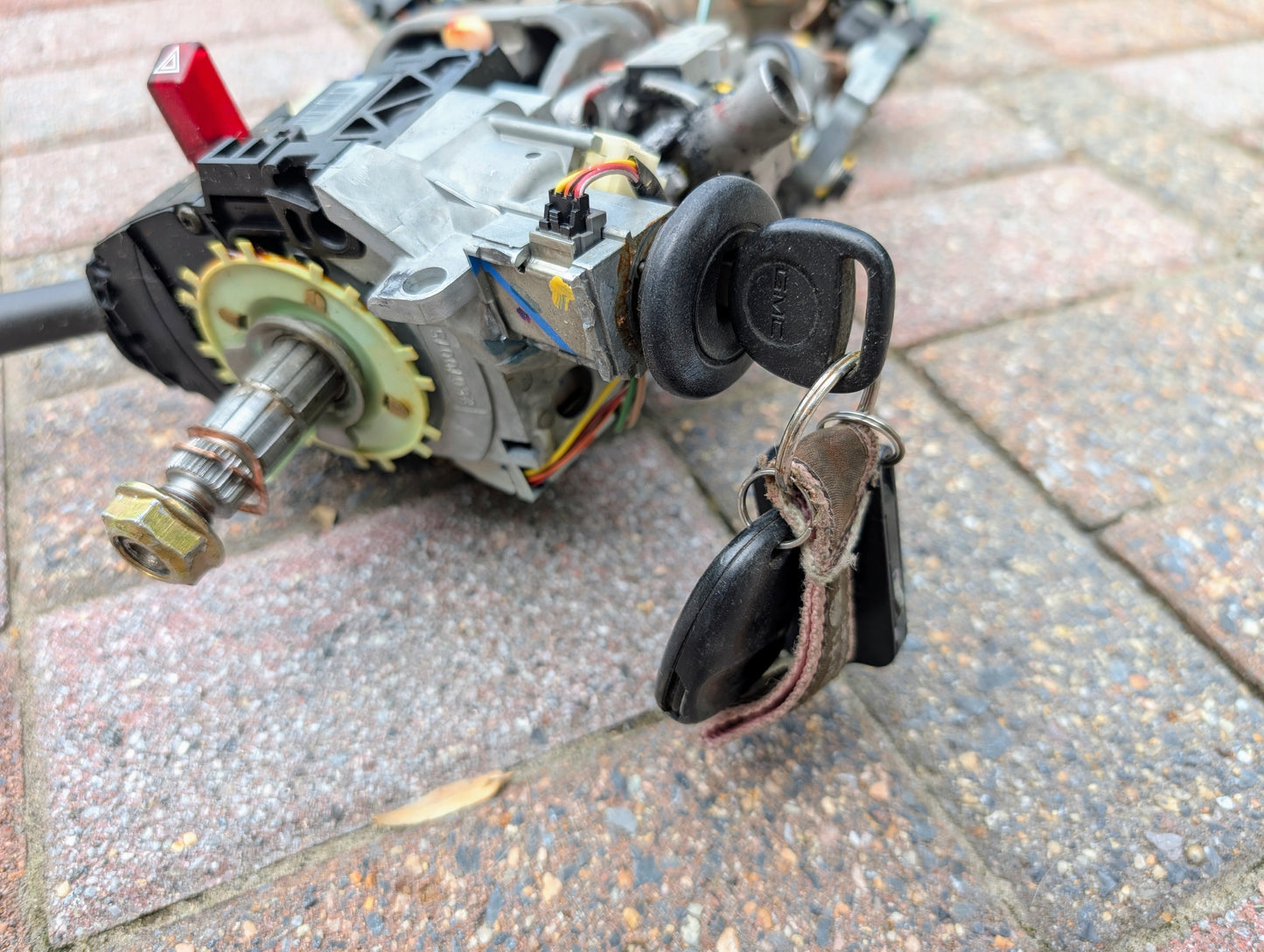 A close-up of a General Motors OEM Steering Column w/KEY for 2000-2002 GM Full Size Trucks shows the ignition switch assembly with keys inserted, revealing wiring and mechanical parts, lying on brick pavement.