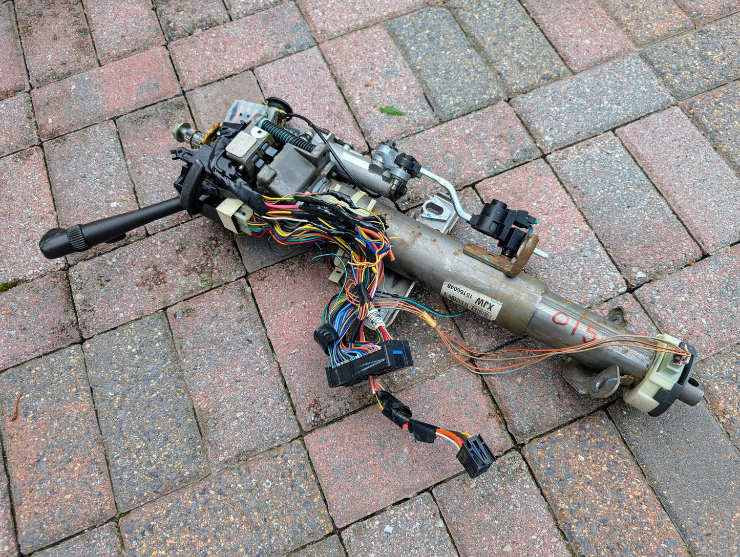A General Motors OEM Steering Column for 2000-2002 GM Full Size Trucks, designed for automatic models, with exposed wiring and switches, lies on brick-paved ground. Electronic parts and connectors are visible on the metal assembly.